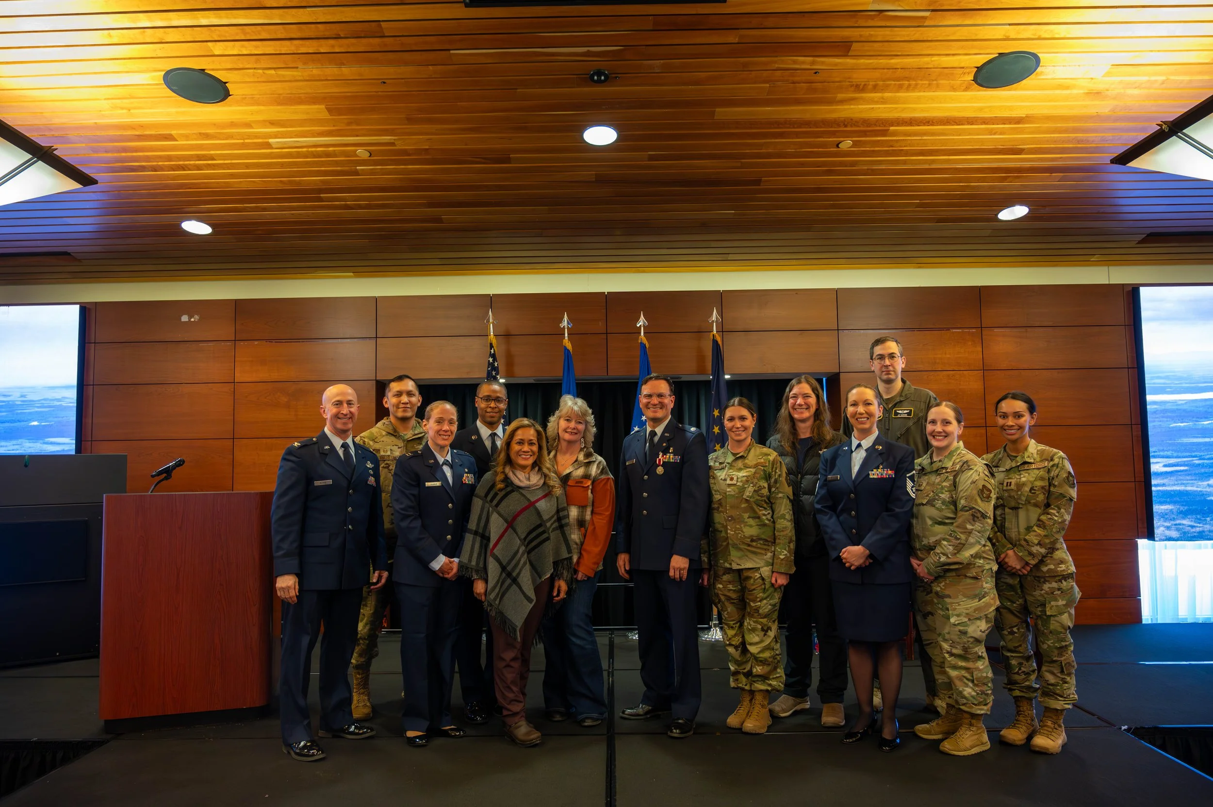 Group of military personnel and civilians standing together in a conference room with wooden walls and flags in the background.