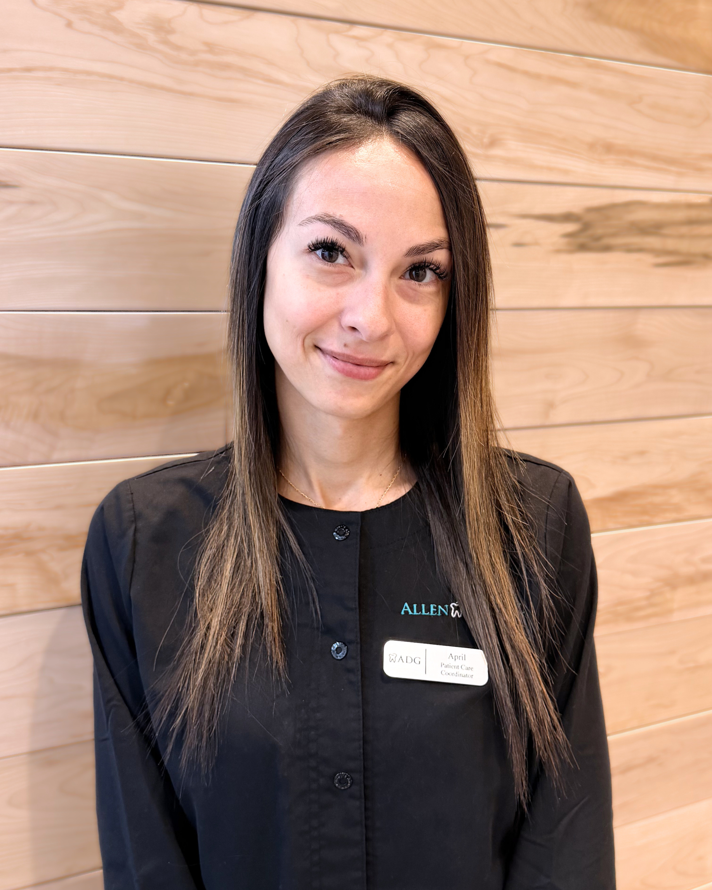 A woman with long brown hair and light skin smiling, standing in front of a wooden wall. She is wearing a black uniform with a name tag that reads 'April' and a badge indicating she is a Patient Care Coordinator at Allen.