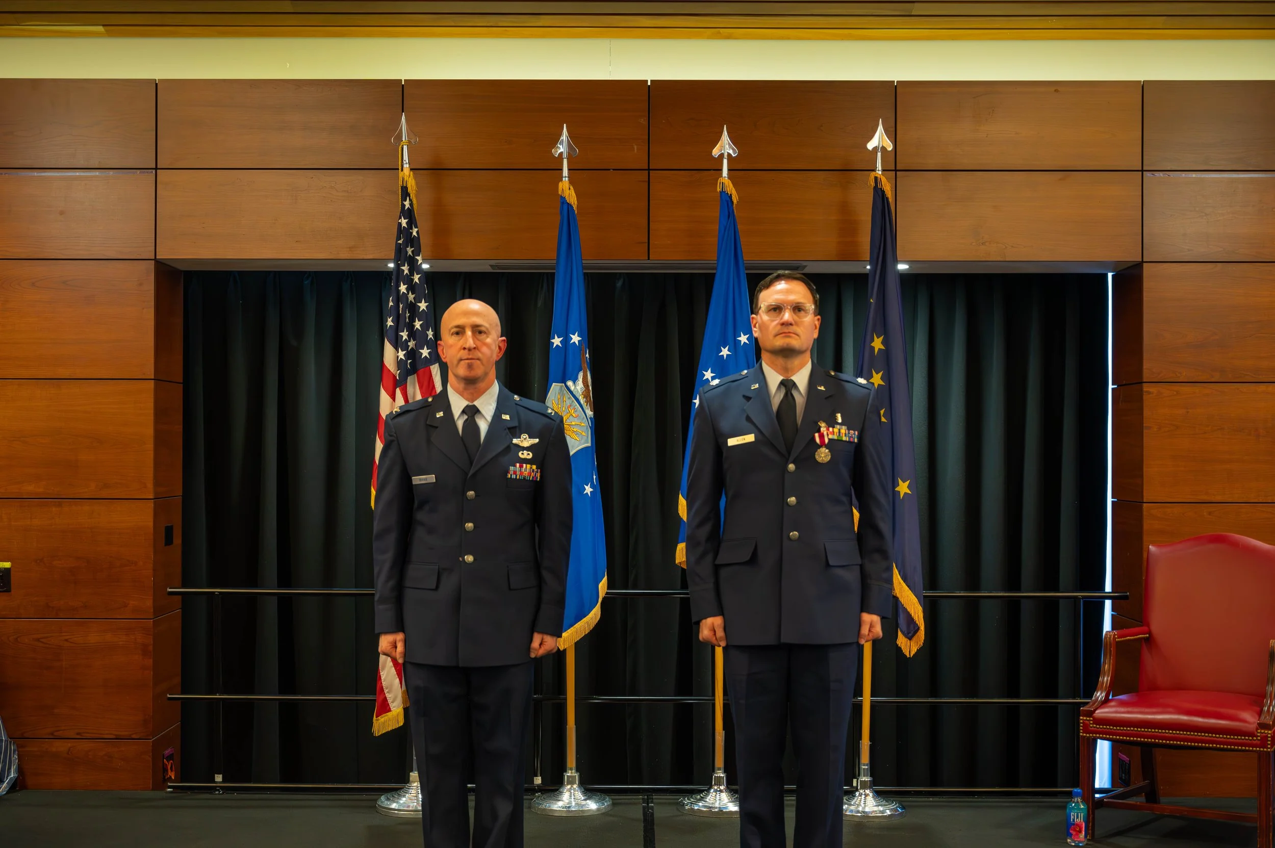 Two men in military uniforms standing in front of a row of flags, including the United States flag, in a room with wooden wall panels and black curtains.
