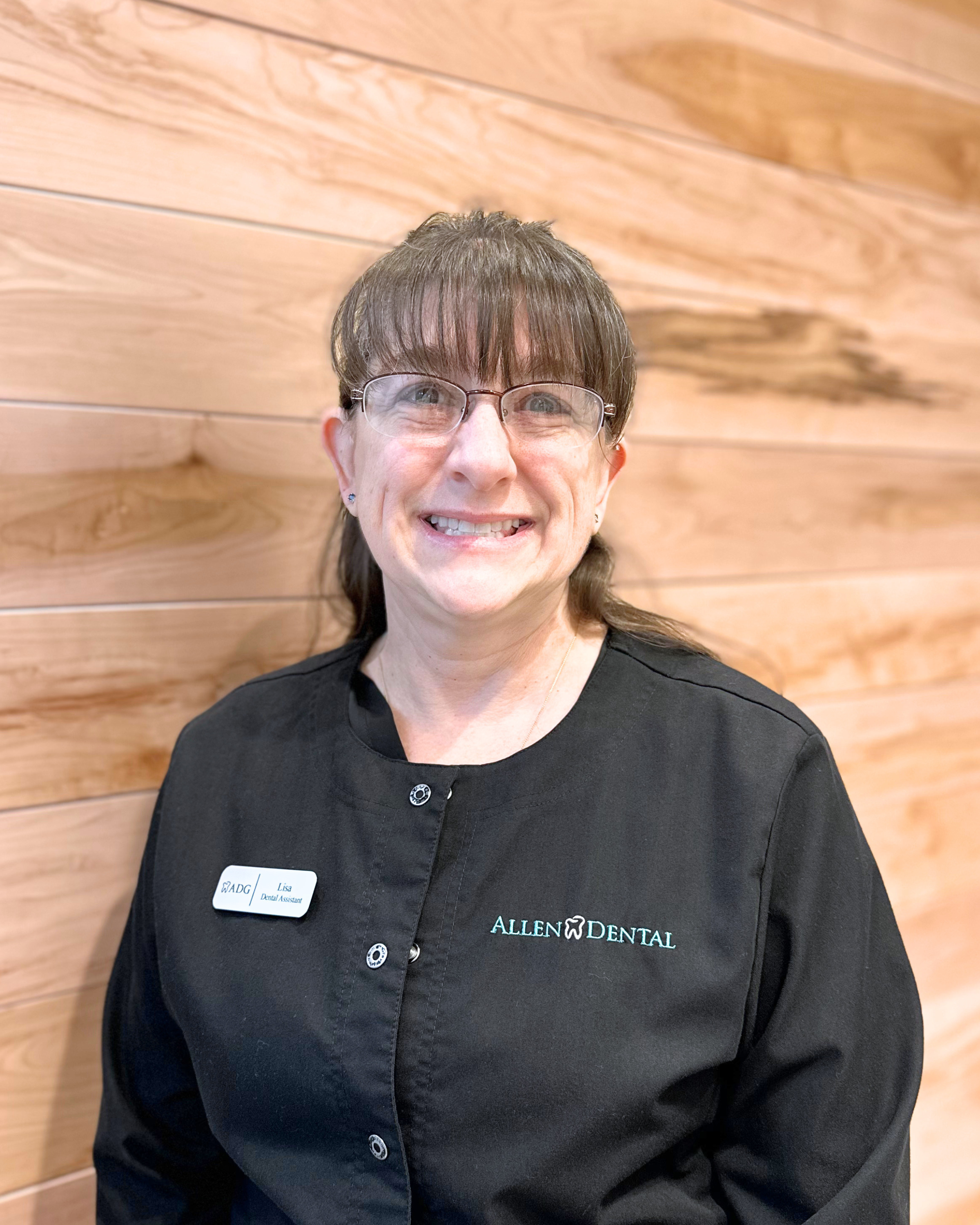 Close-up of a smiling woman with brown hair and glasses, wearing a black uniform with a name tag that reads 'Lisa, Dental Assistant' and the logo 'Allen Dental' on it, standing in front of a wooden wall.