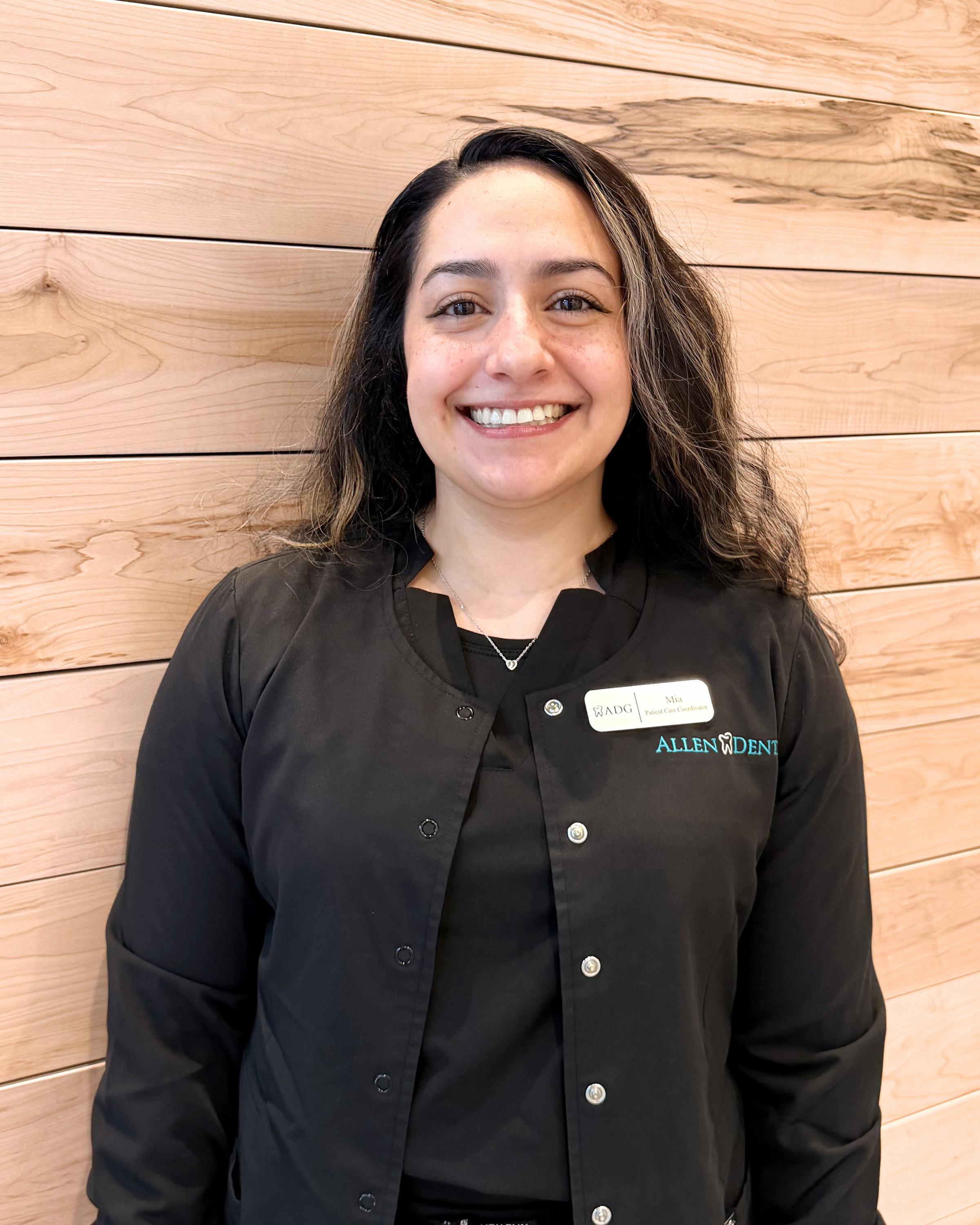 A woman with long curly hair and light skin wearing a black professional jacket with a name tag, smiling, standing in front of a wooden wall.