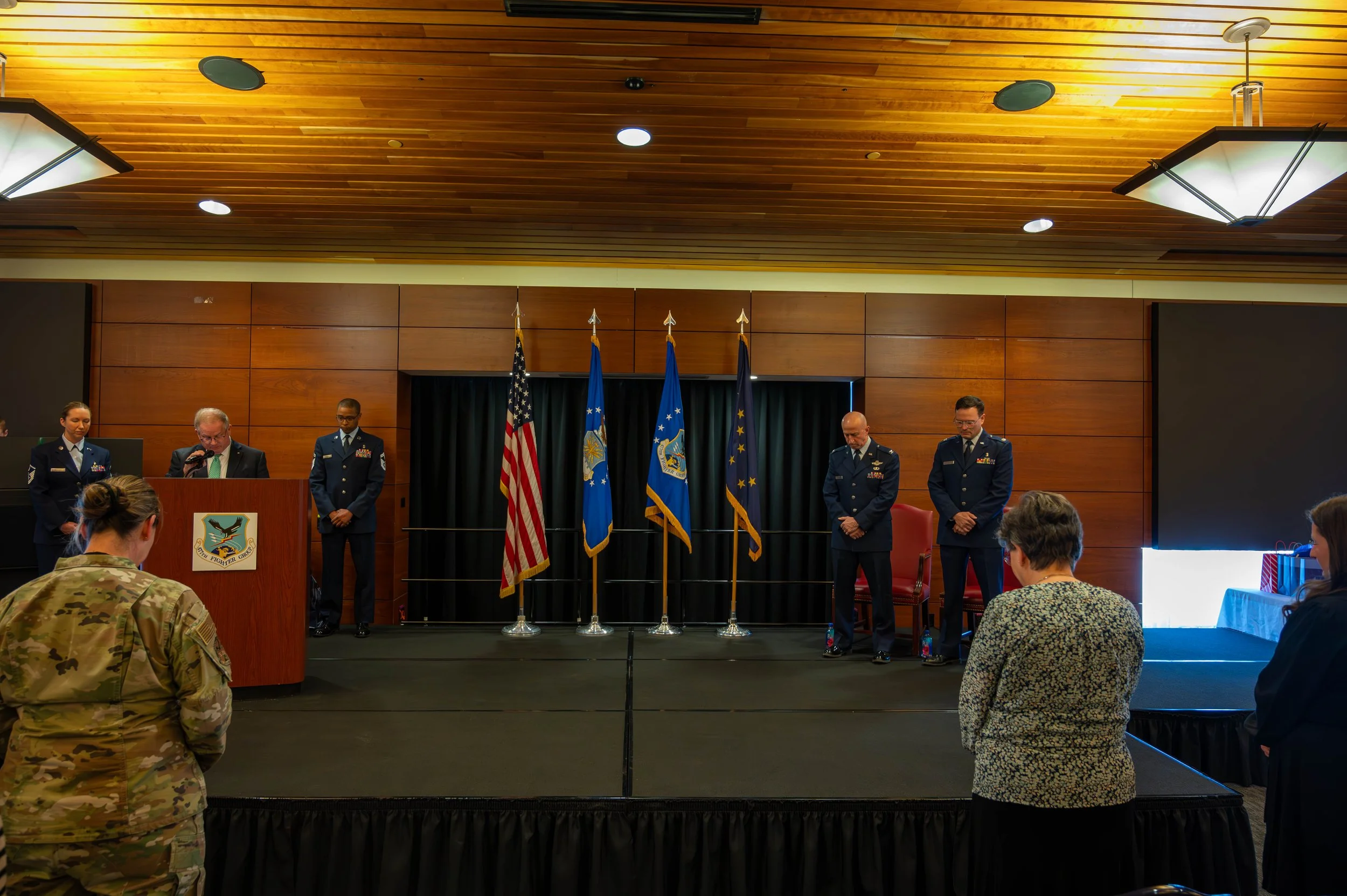 Military personnel and officials standing on stage during a formal event with American and military flags.