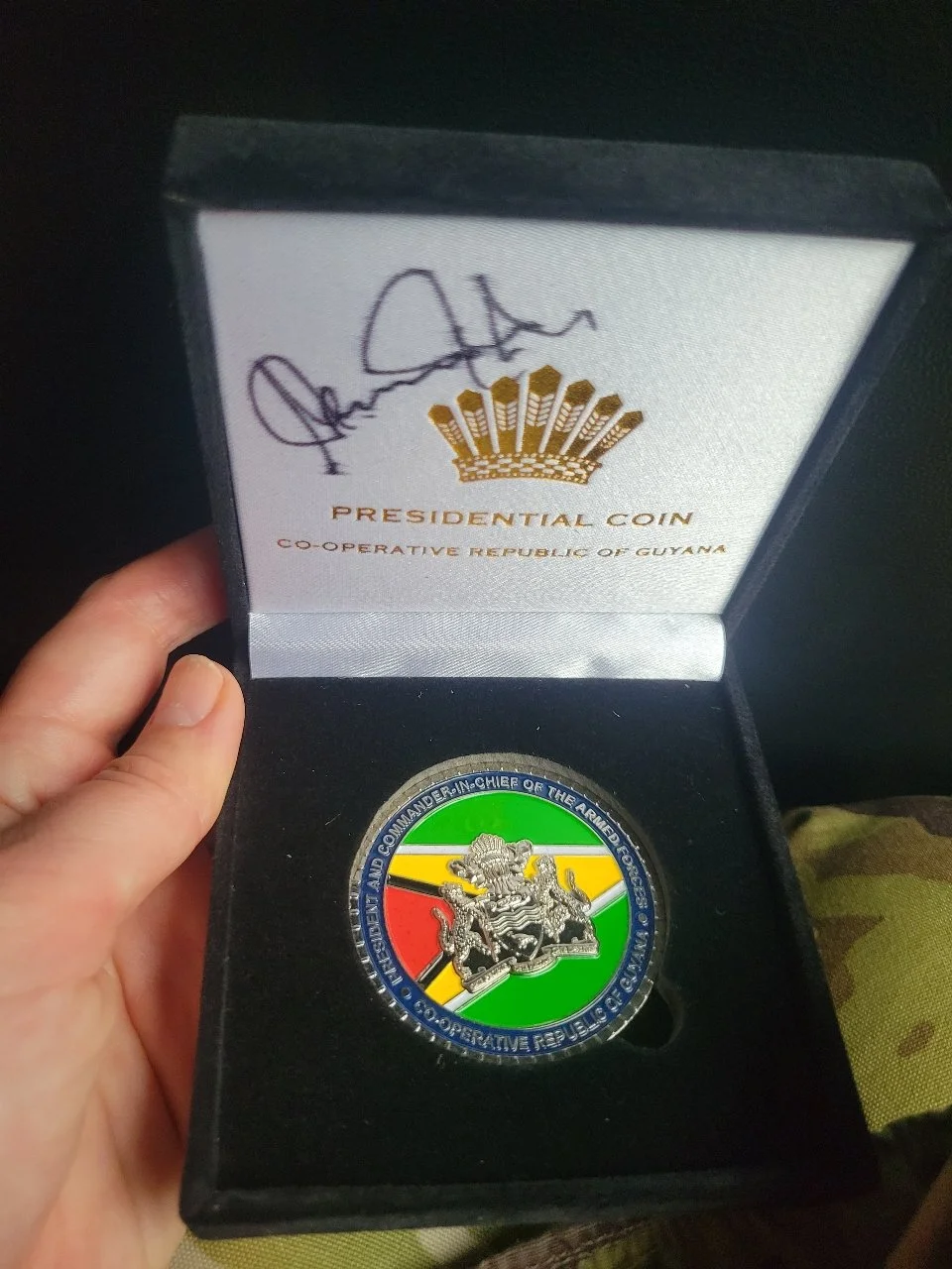 A hand holding an open box featuring a coin with colorful background and a lion emblem. The box interior has a signature and the words "Presidential Coin" and "Co-Operative Republic of Guyana."