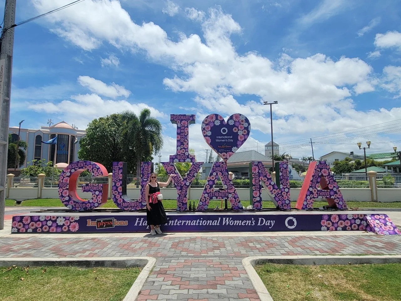 A woman in a black dress with sunglasses standing next to large floral-decorated letters spelling 'GREAT' and 'LOVE,' with a purple sign indicating International Women's Day, in front of a green park with buildings and blue sky with clouds.
