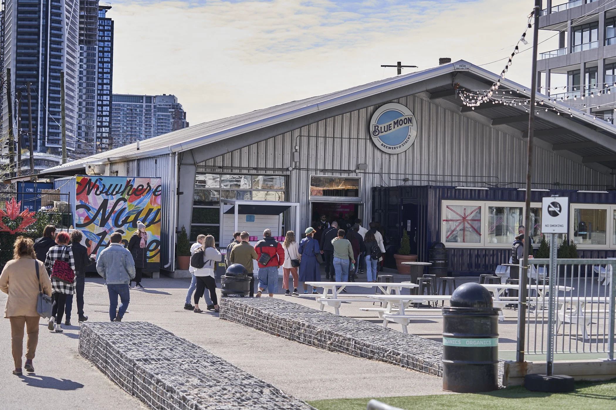 People line up outside a brewery called Blue Moon during daytime, in an urban area with high-rise buildings. The brewery has a metal building with a sign and string lights hanging above.
