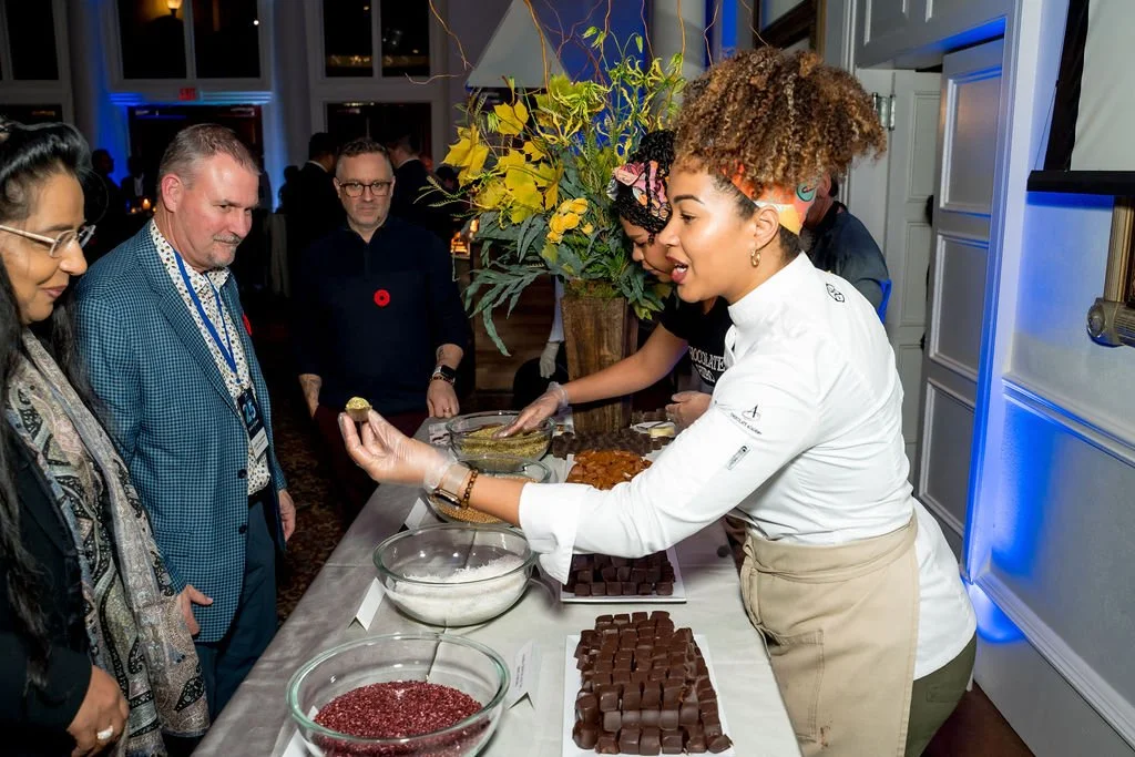 A woman in a white chef's coat demonstrating chocolate confections at a dessert table, with several people watching her in a decorated indoor event space.