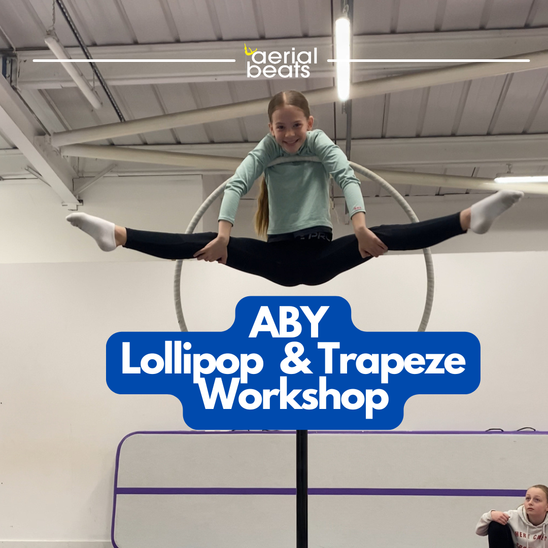 A young girl performing an aerial split on a lyra ring hang in a gym, smiling at the camera with a sign over her that reads 'ABY Lollipop & Trapeze Workshop.'