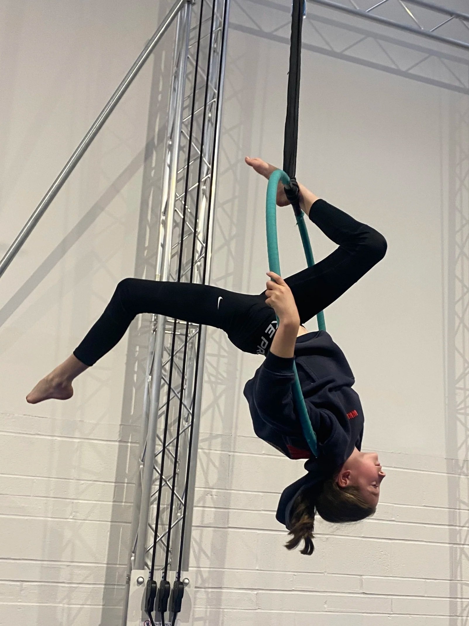 A young girl performing a backbend on gymnastic rings in an indoor gym, with a metal structure in the background.