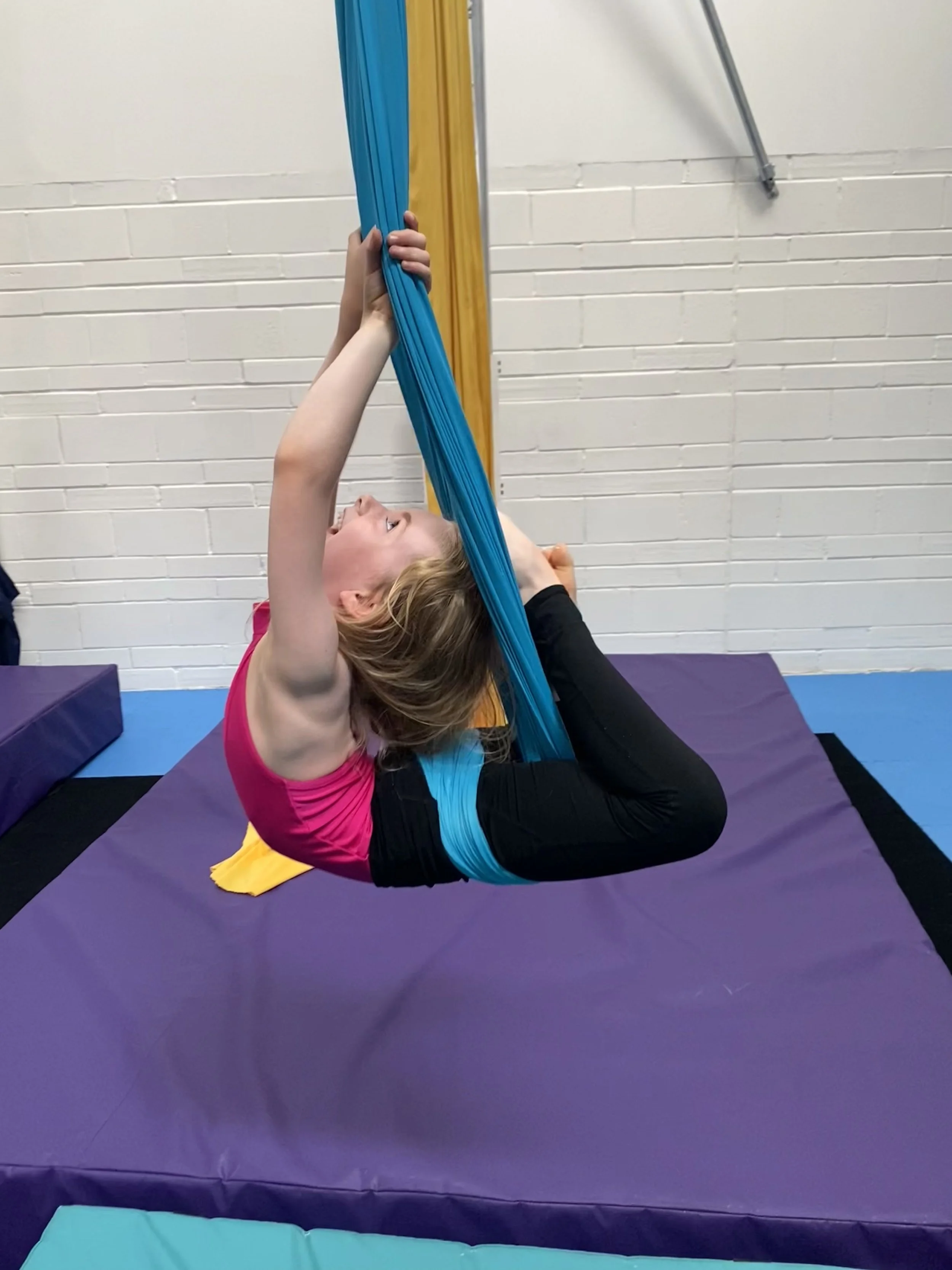 A young girl with blonde hair practicing aerial silks in a gym, lying upside down with her legs wrapped around blue fabric, held by her hands, on purple padded flooring.