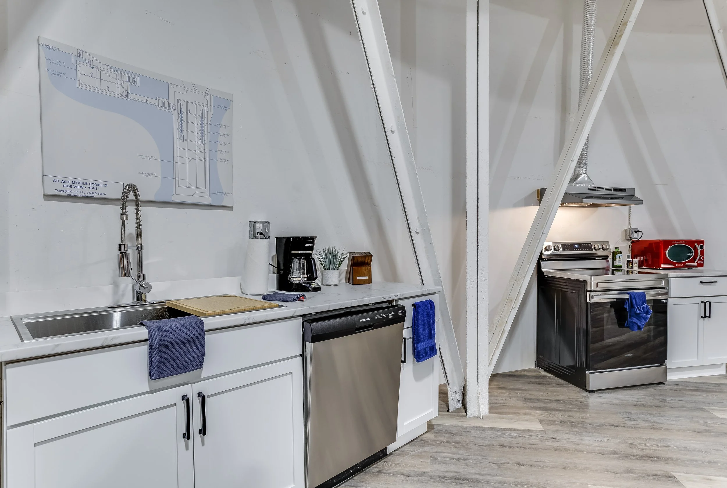 A modern kitchen with white cabinets, stainless steel sink, black dishwasher, coffee maker, potted plant, and kitchen utensils on a white countertop. There is a blueprint on the wall and a stainless steel range hood above the stove. The floor is wood