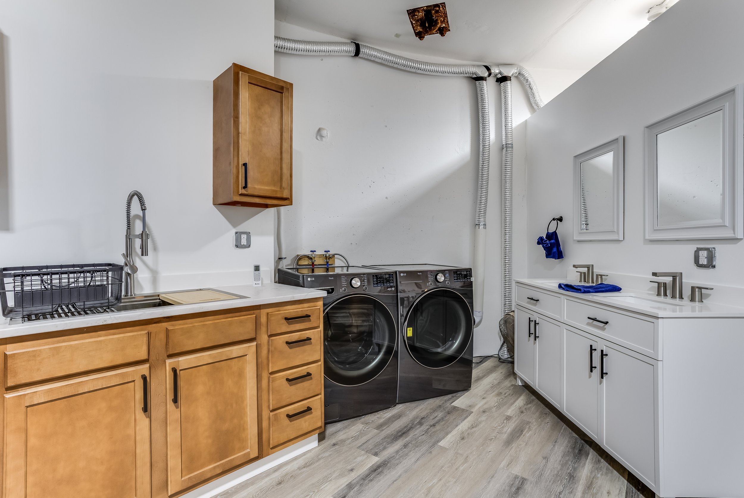 Laundry room with wooden cabinets, a stainless steel sink, a drying rack, and a washing machine and dryer. The wall has ducting and mirrors, with a towel hanging near the sink.