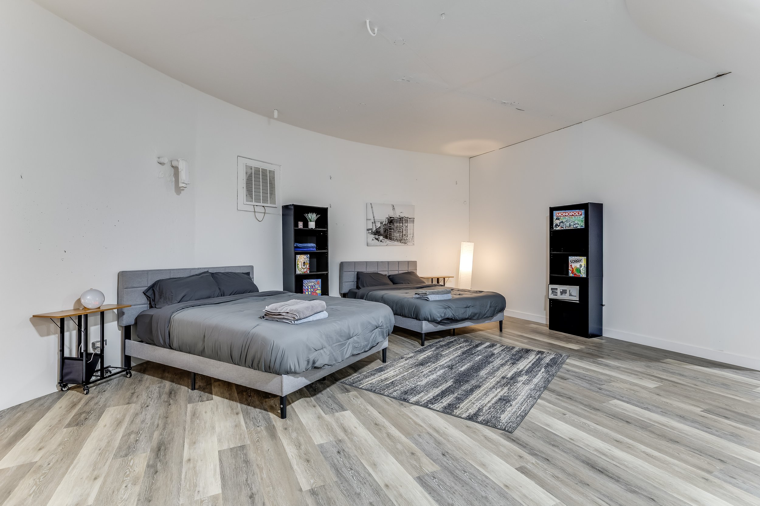 A bedroom with two beds, pillows, and folded towels. There are black bookshelves with books and decorative items, a small table with a lamp, a floor lamp, and wall art. The flooring is light wood, and the ceiling has visible damage.