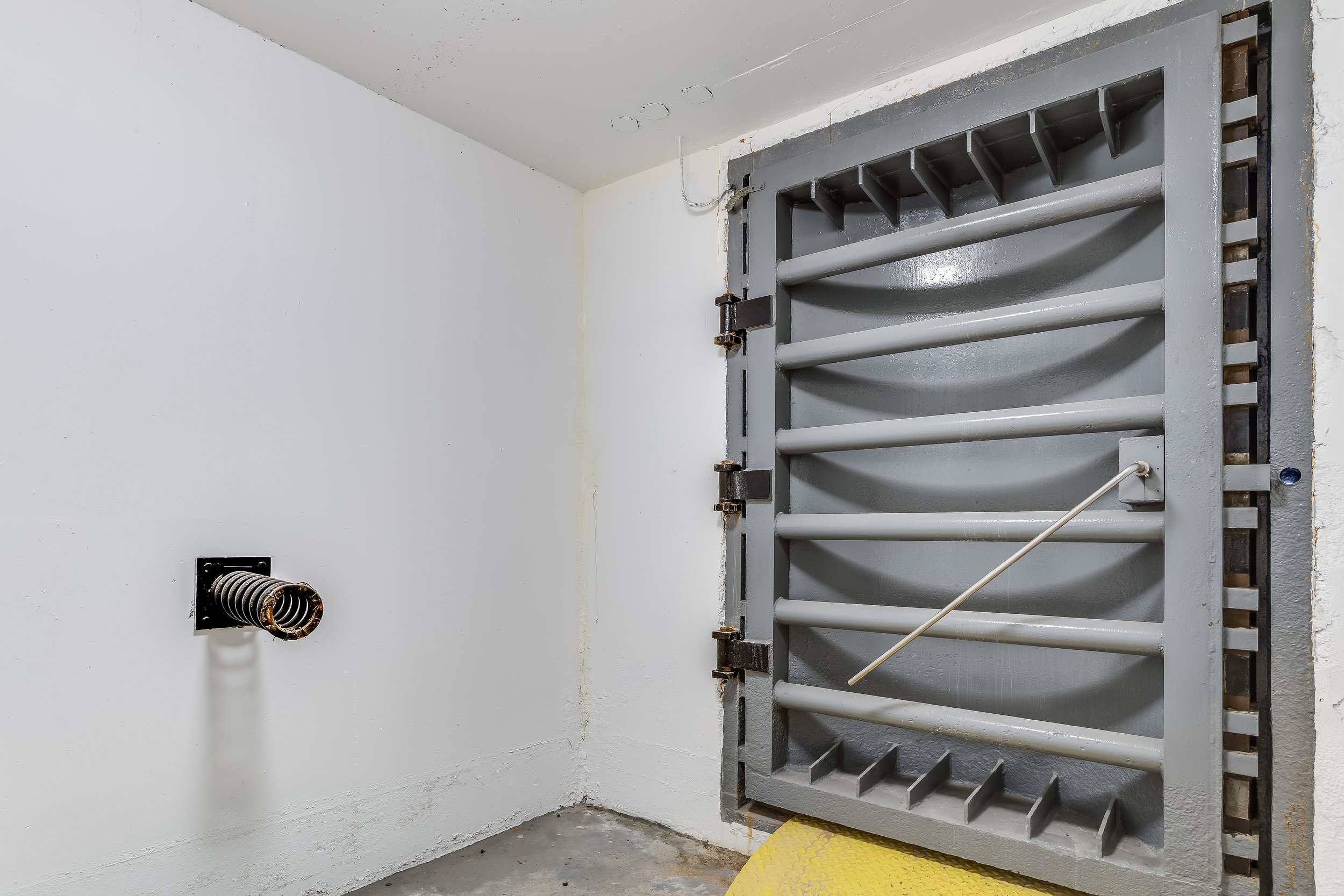 Interior of a prison cell with gray metal bunk beds, a yellow safety mat, a vent pipe on the white wall, and cracked ceiling.