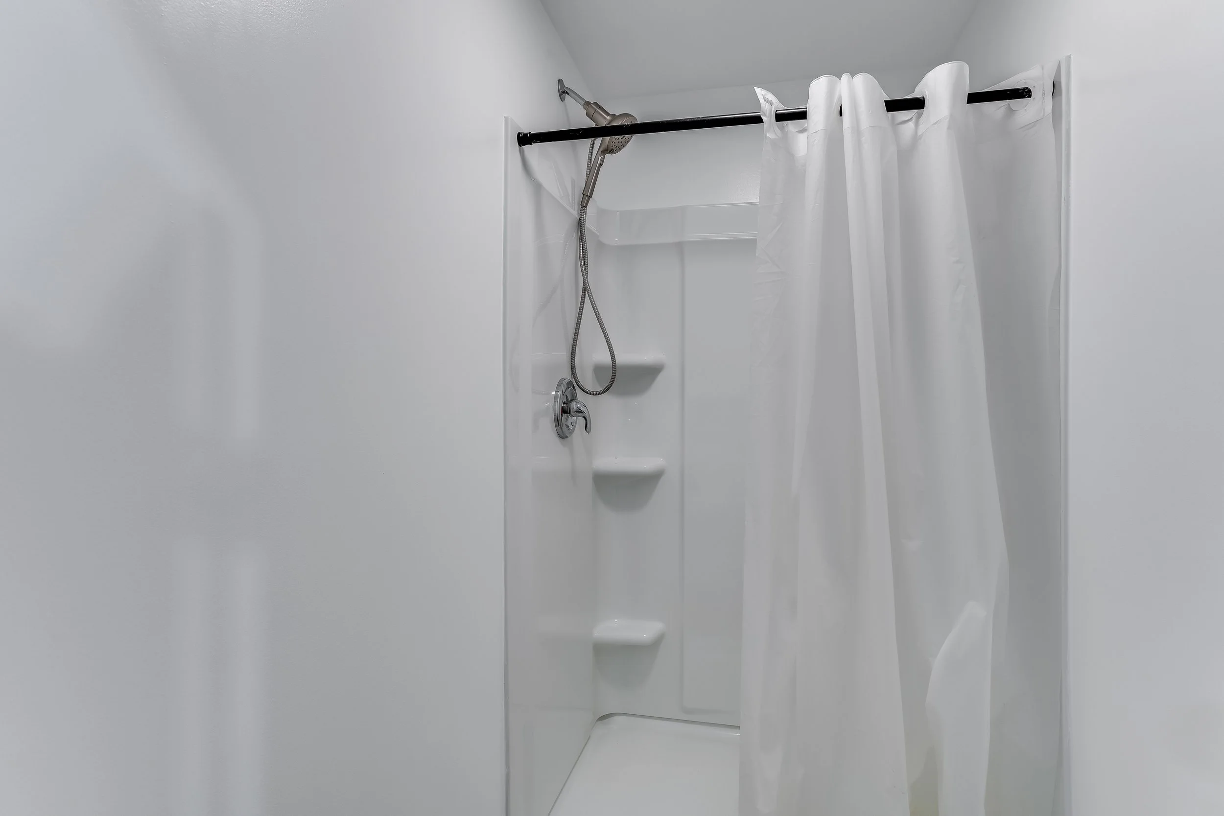 Empty white shower stall with soap dish and a black shower curtain rod, white shower curtain partially hanging.