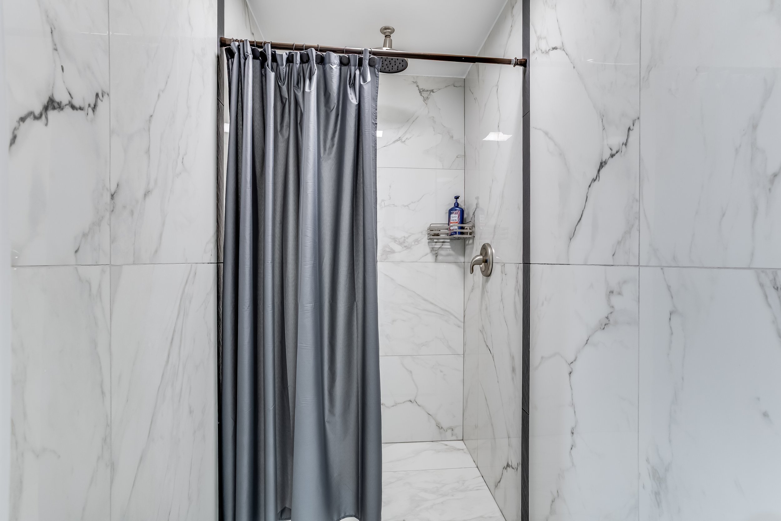 A bathroom shower area with marble wall tiles, a gray shower curtain, a showerhead, and a small shelf holding shower products.