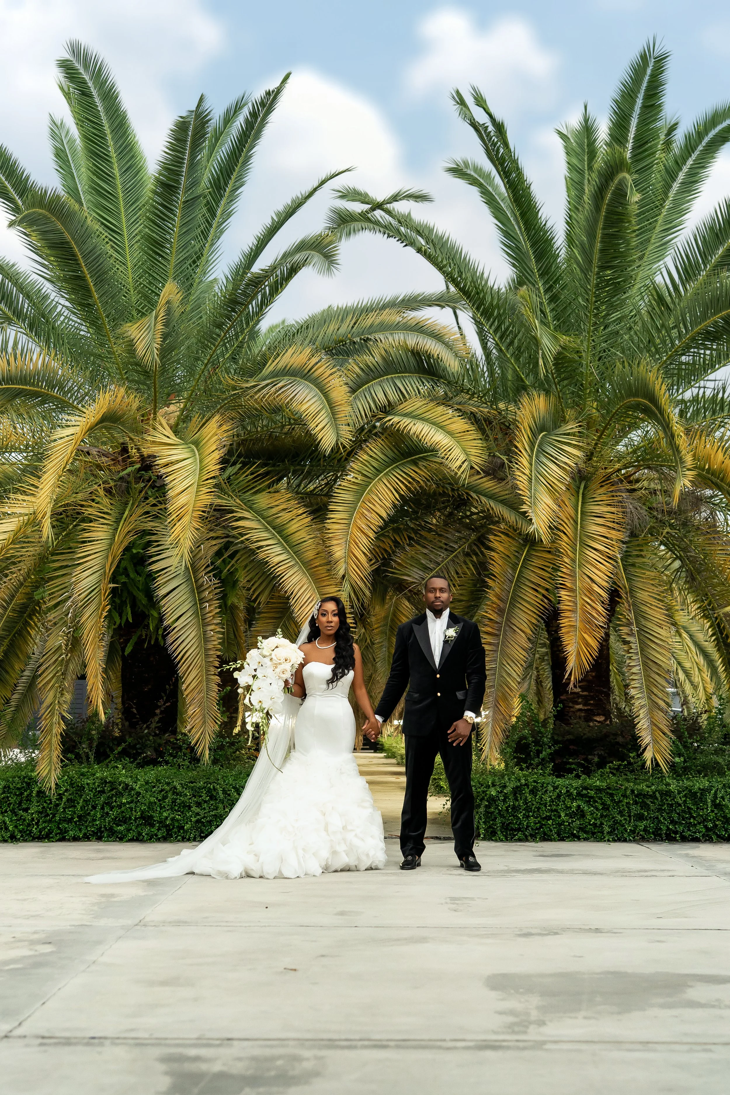 A bride and groom in wedding attire holding hands standing on a paved path in front of tall palm trees with green foliage against a cloudy sky.