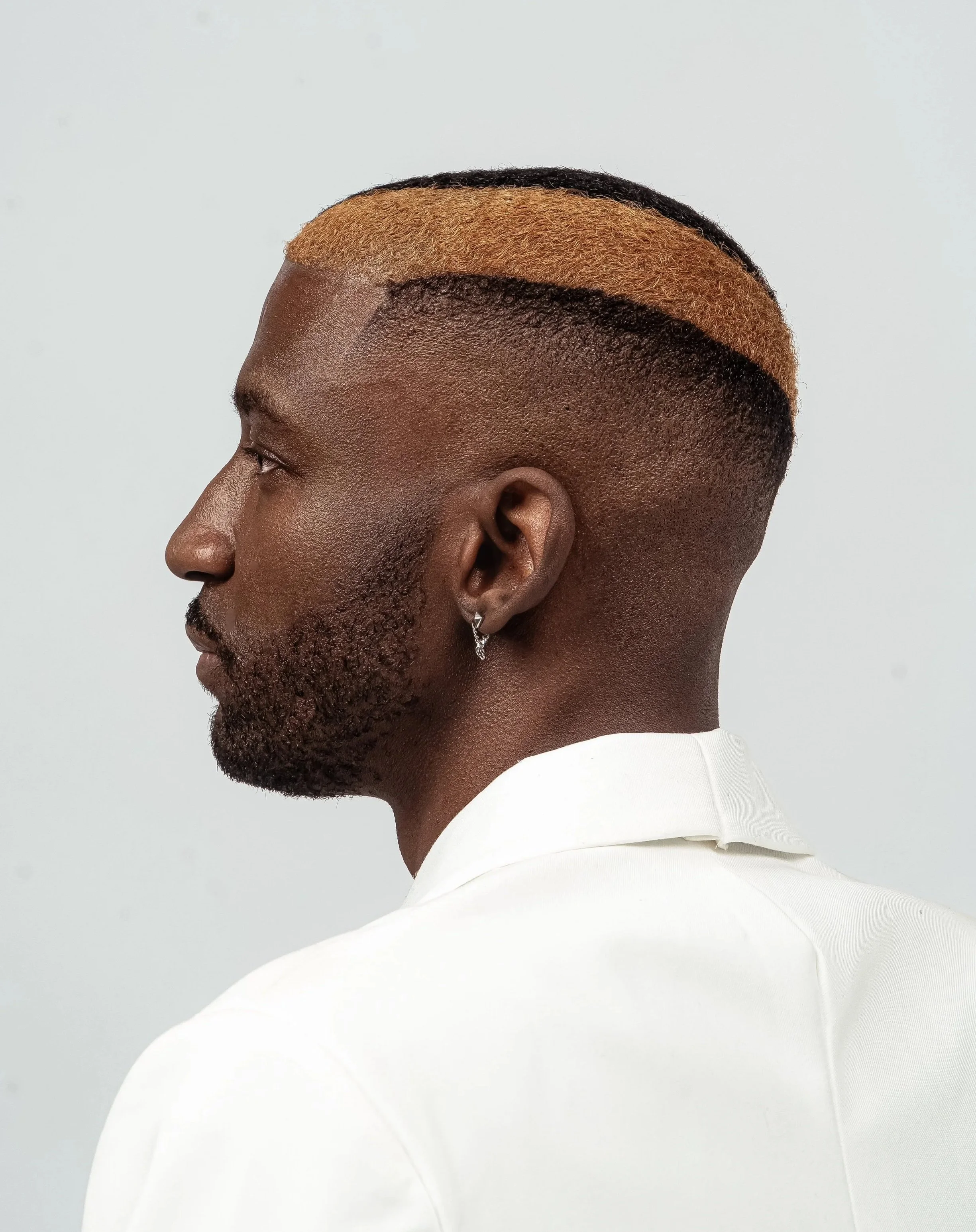 A man with a dark skin tone, short hair with a blonde section, wearing a small hoop earring, dressed in a white shirt, and looking to the left against a plain light gray background.