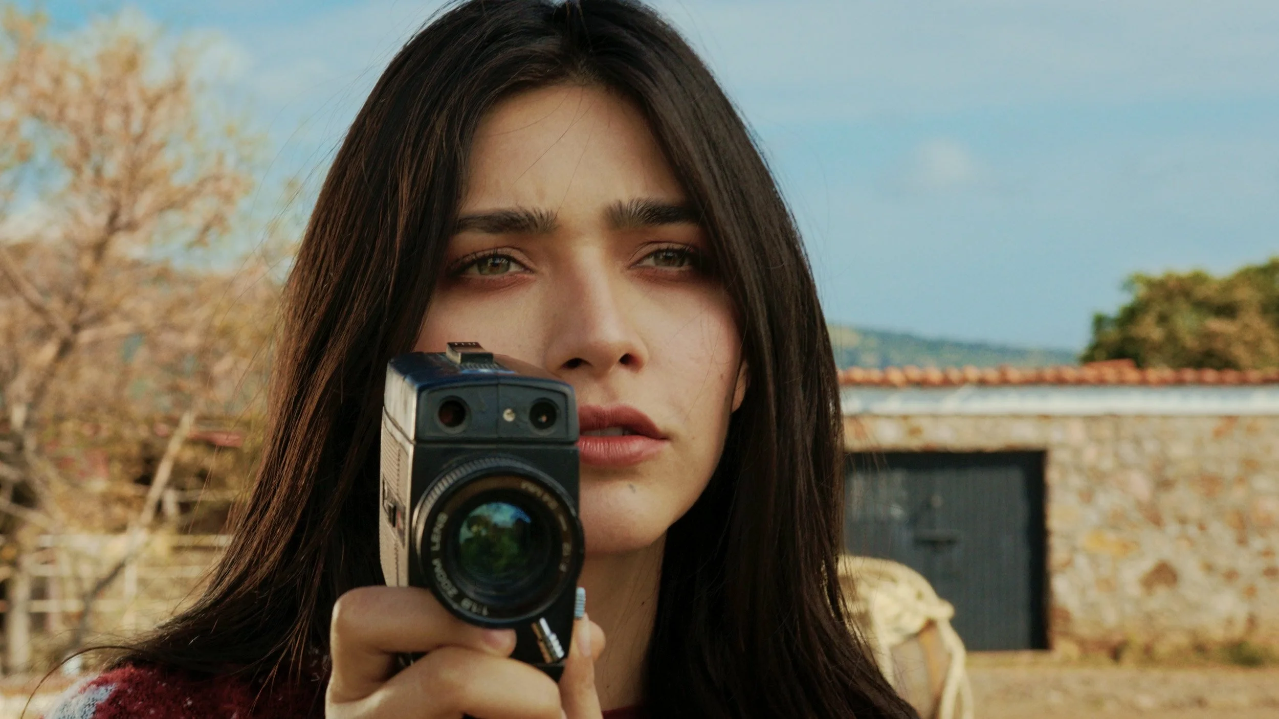 A woman with long dark hair holding a vintage camera close to her face, outdoors with trees and a stone building in the background.
