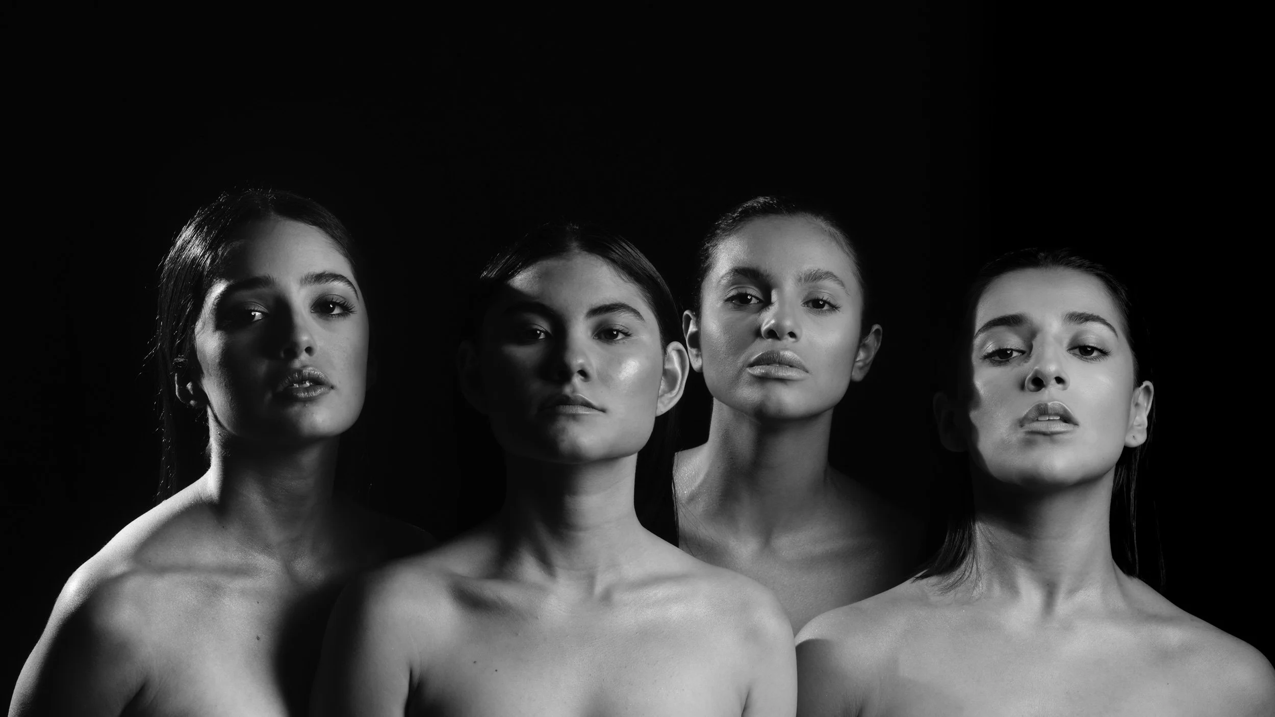 Black and white portrait of four topless women with serious expressions, standing close together against a dark background.