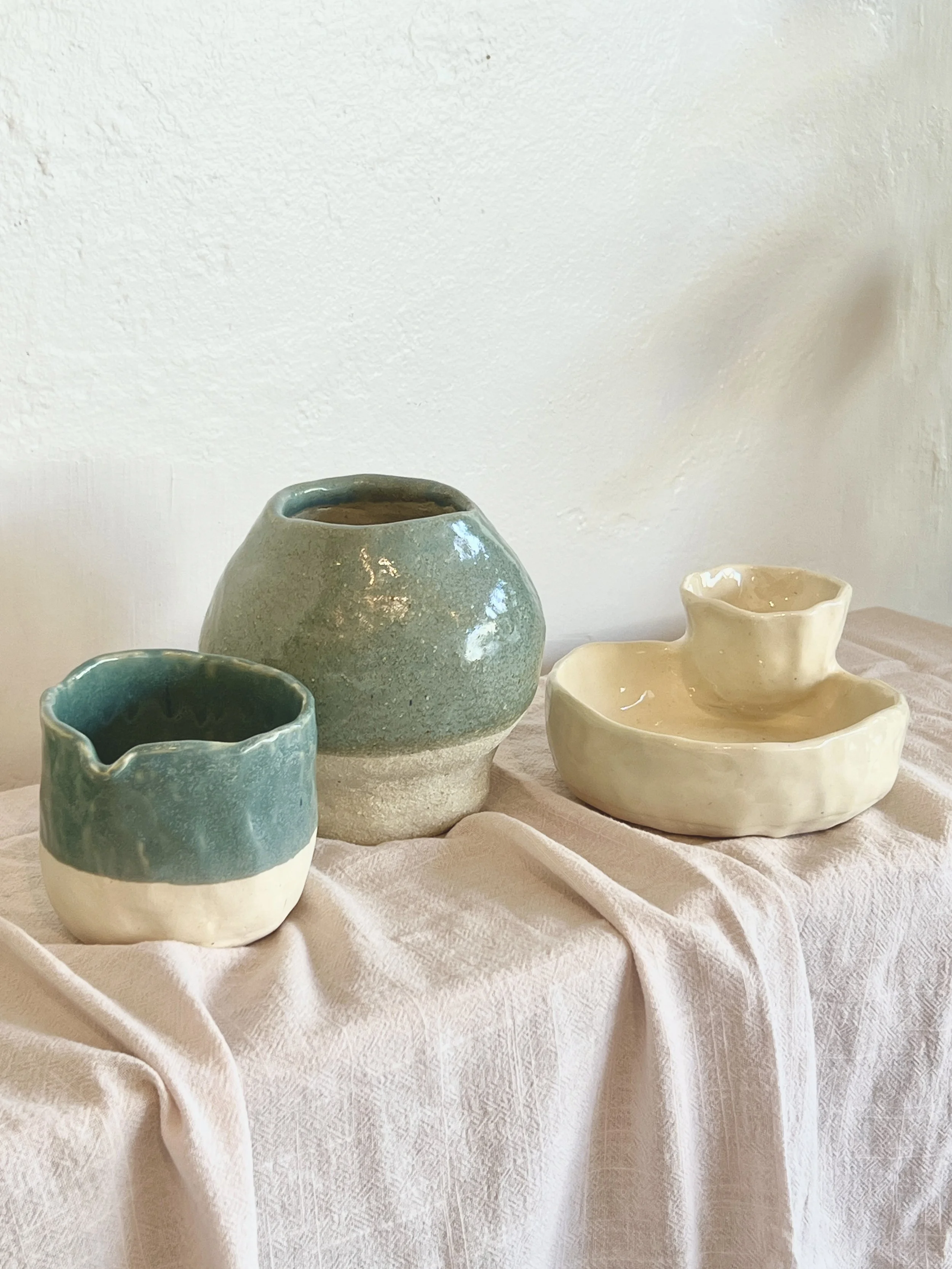 Three ceramic pottery pieces on a draped light-colored fabric tablecloth against a plain wall.