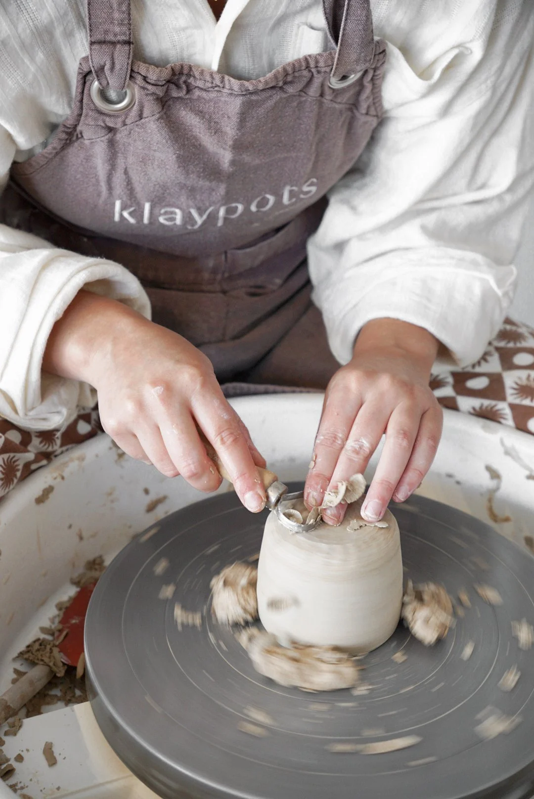 Person wearing an apron labeled 'klaypots' working on a pottery wheel, shaping a clay pot with fingers while the wheel spins.