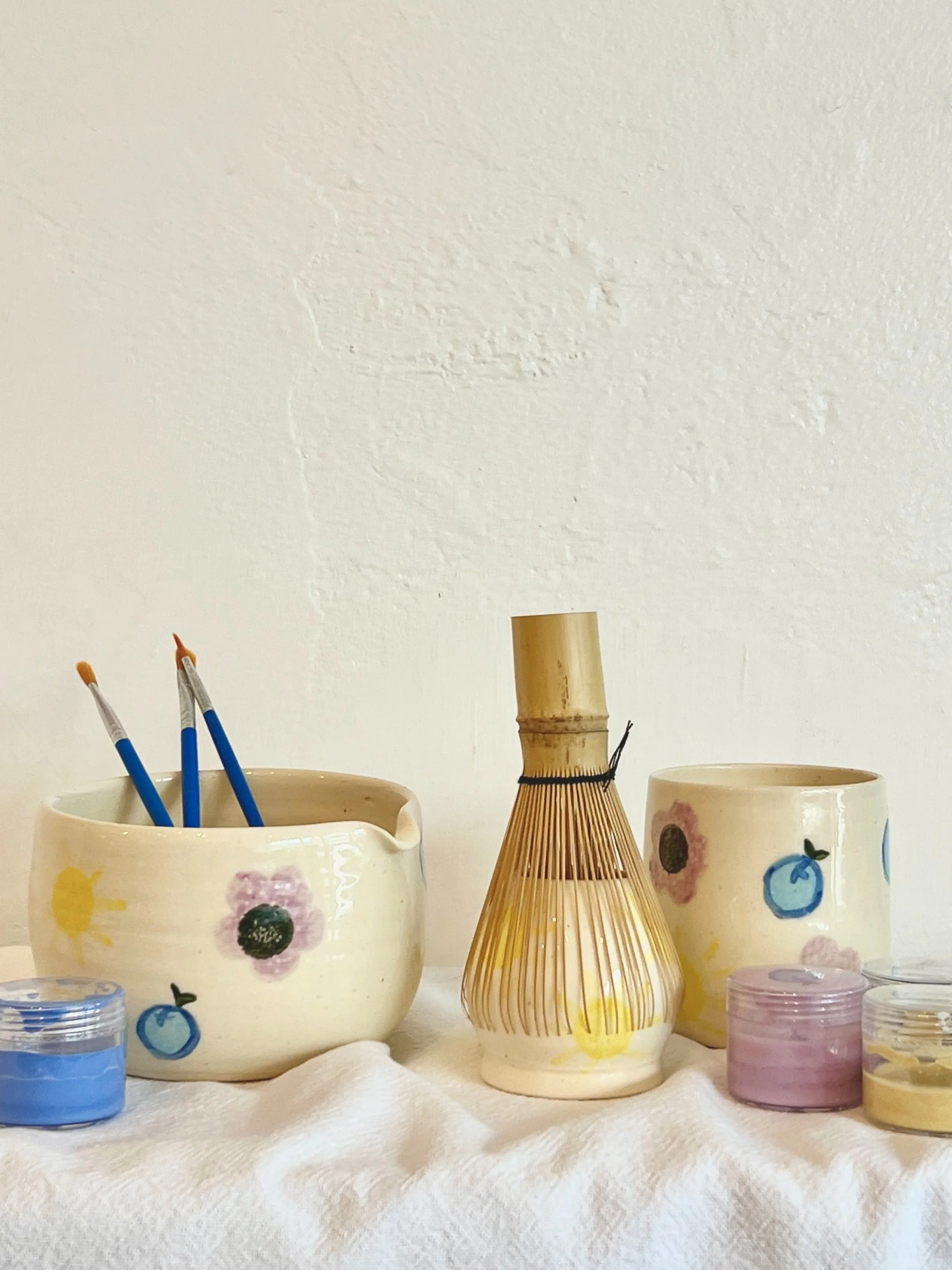 Decorative ceramics with painted fruit and sunflower designs, including a rice paddle-shaped container holding paintbrushes, and small containers of paint on a white fabric surface.