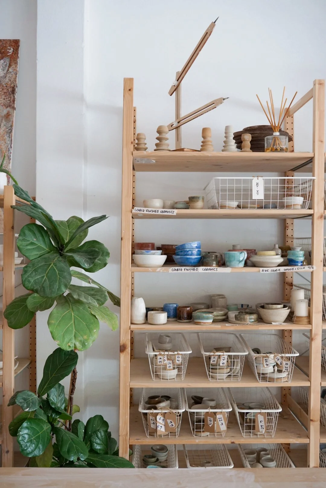Wooden shelf displaying various finished ceramics bowls, cups, and plates in baskets, with labels, next to a large green plant.