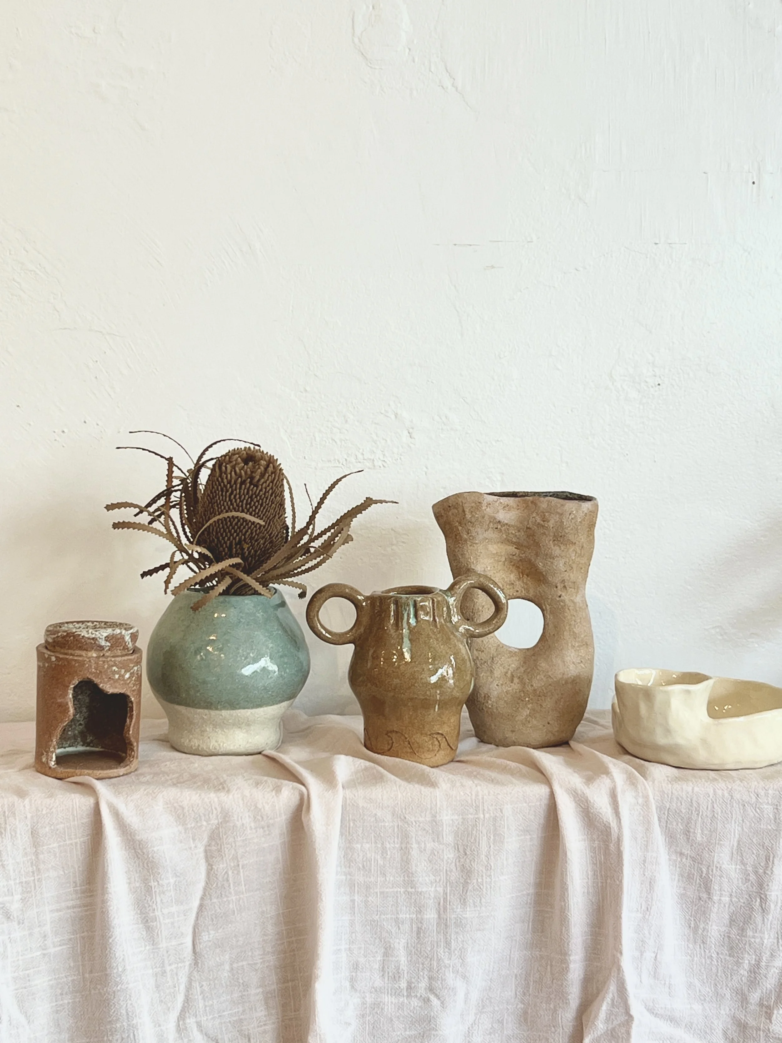 A collection of ceramic vases and sculptures on a light-colored, textured tablecloth against a plain white wall.