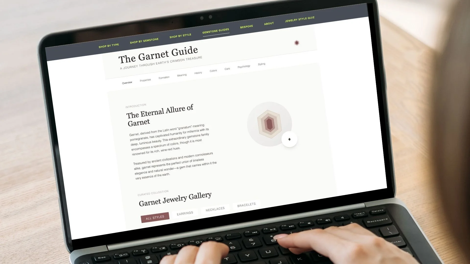 A person browsing a website about garnets on a laptop. The website is titled 'The Garnet Guide' and features information about garnet gemstones, including a color diagram and a garnet jewelry gallery.