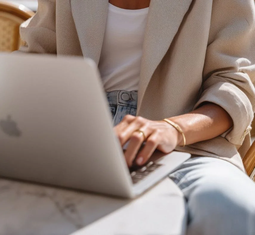 Cropped lifestyle photo of a person in a beige blazer and white top typing on a laptop while seated, wearing a simple gold bracelet and ring. Soft, warm lighting with a neutral background.