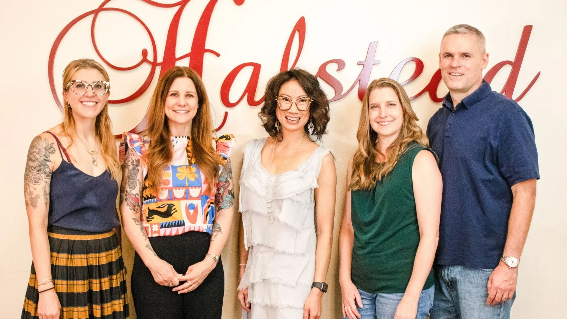 Group of five women and one man standing in front of a wall with red cursive text that says 'Fasted'. The women have various hair colors and styles, and the man has short gray hair. They are smiling and dressed in casual clothing.