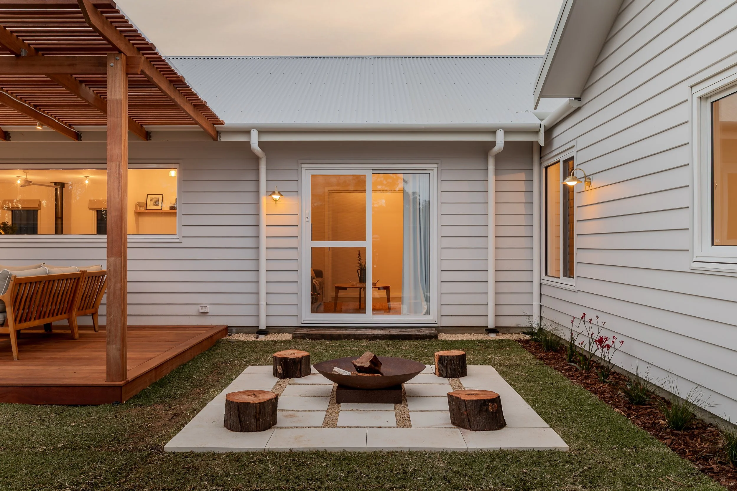 The image shows the exterior of a modern house with white siding, large glass sliding door, and outdoor lighting. There is a wooden deck with outdoor furniture, and a small paved area with tree stumps arranged around a fire pit. The sky appears cloud