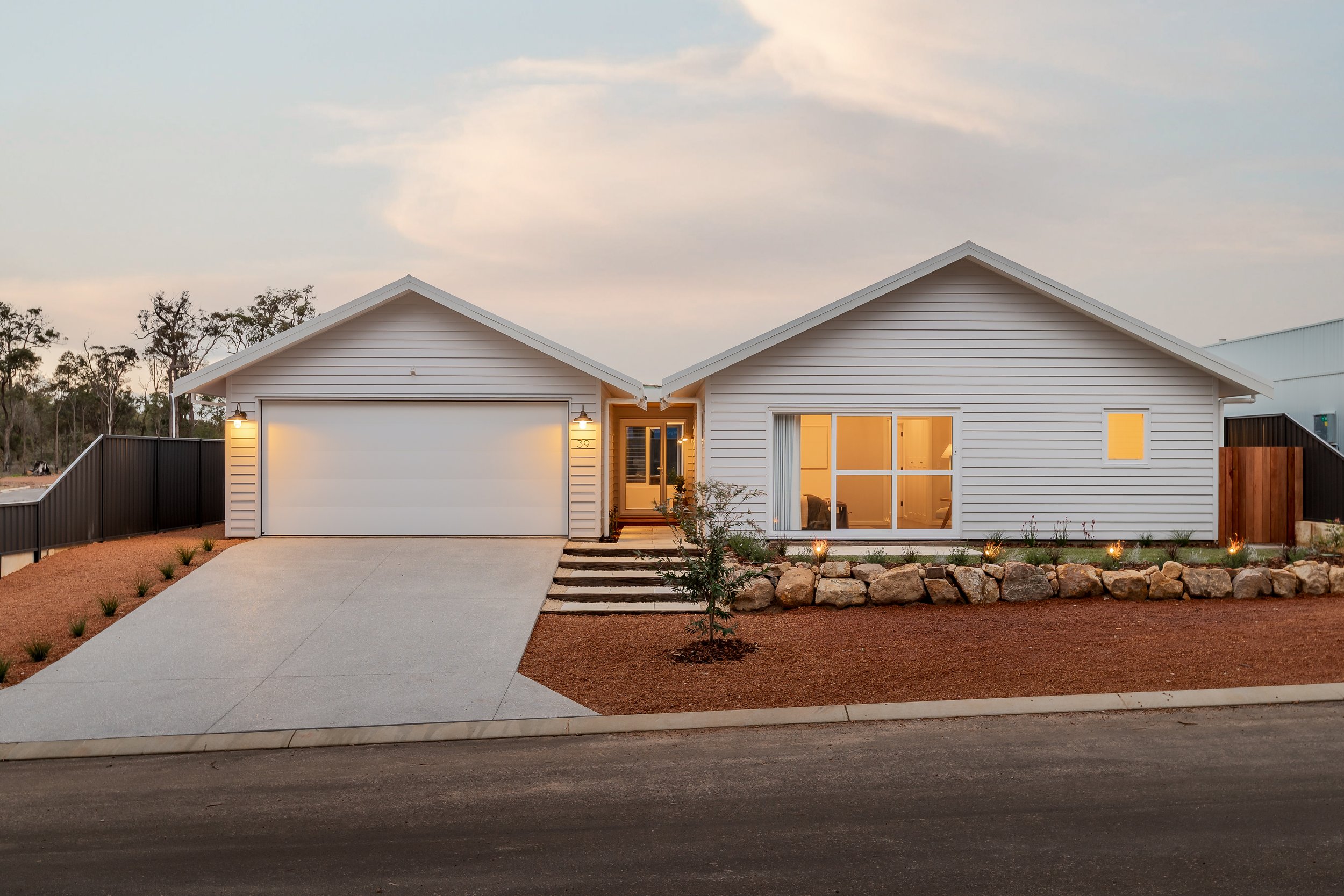 Margaret River, Western Australia. A modern white house with a two-car garage, a concrete driveway, a small front yard with rocks and young plants, and warm interior lights visible through large windows, at dusk. Real Estate Photography.