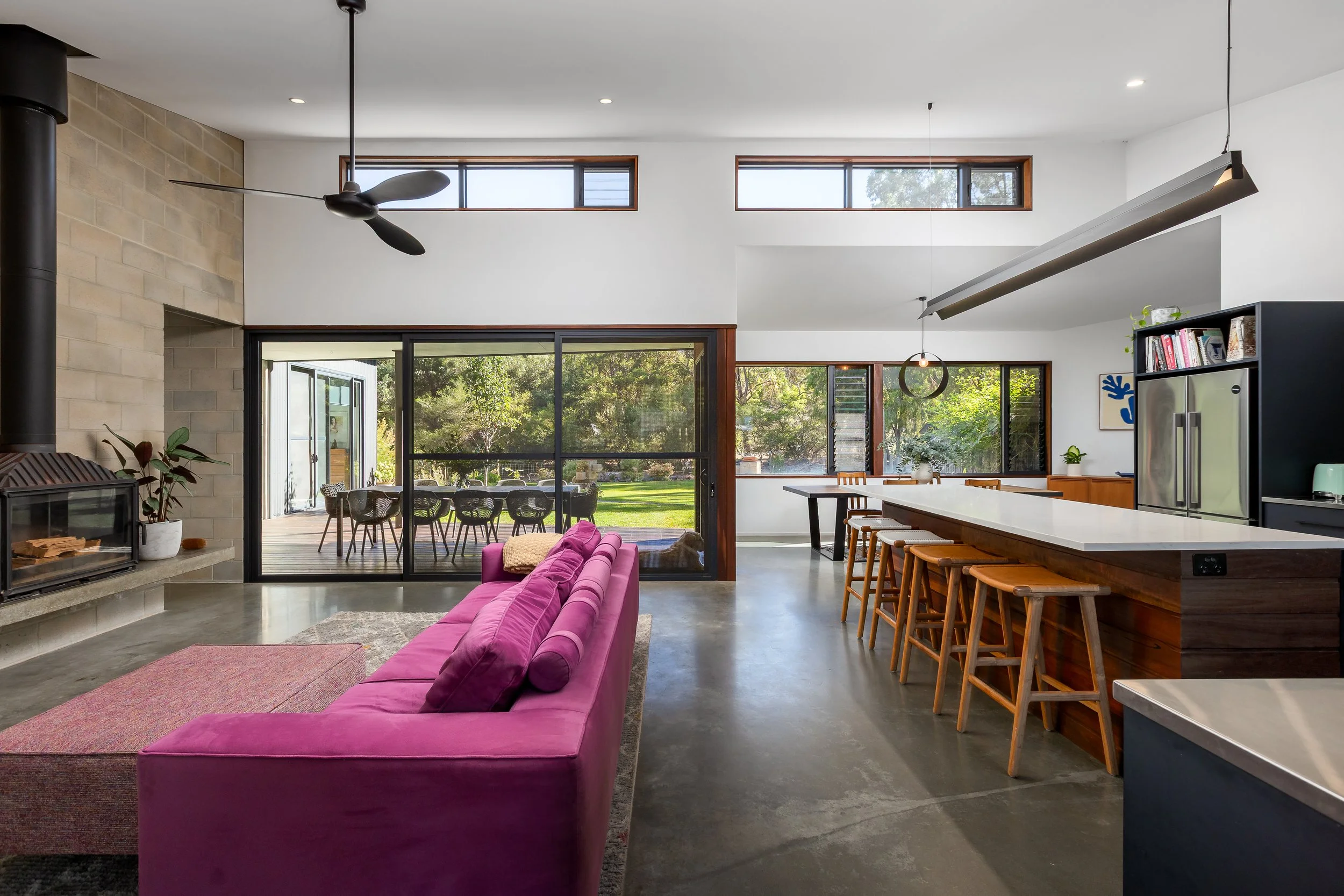 Yallingup, Western Australia. Modern open-concept living space with large windows, pink sectional sofa, wooden dining counter with stools, view of outdoor patio and greenery. Real Estate Photography.