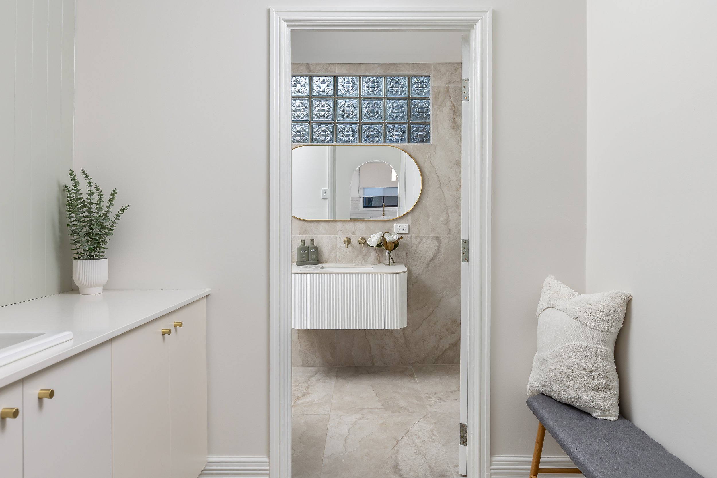 Harvey, Western Australia. A bathroom with a mirror and glass block window, with a small sink area, a decorative soap holder, bottles, and a potted plant to the side. A gray bench with a beige fluffy pillow is to the right. Real Estate Photography.