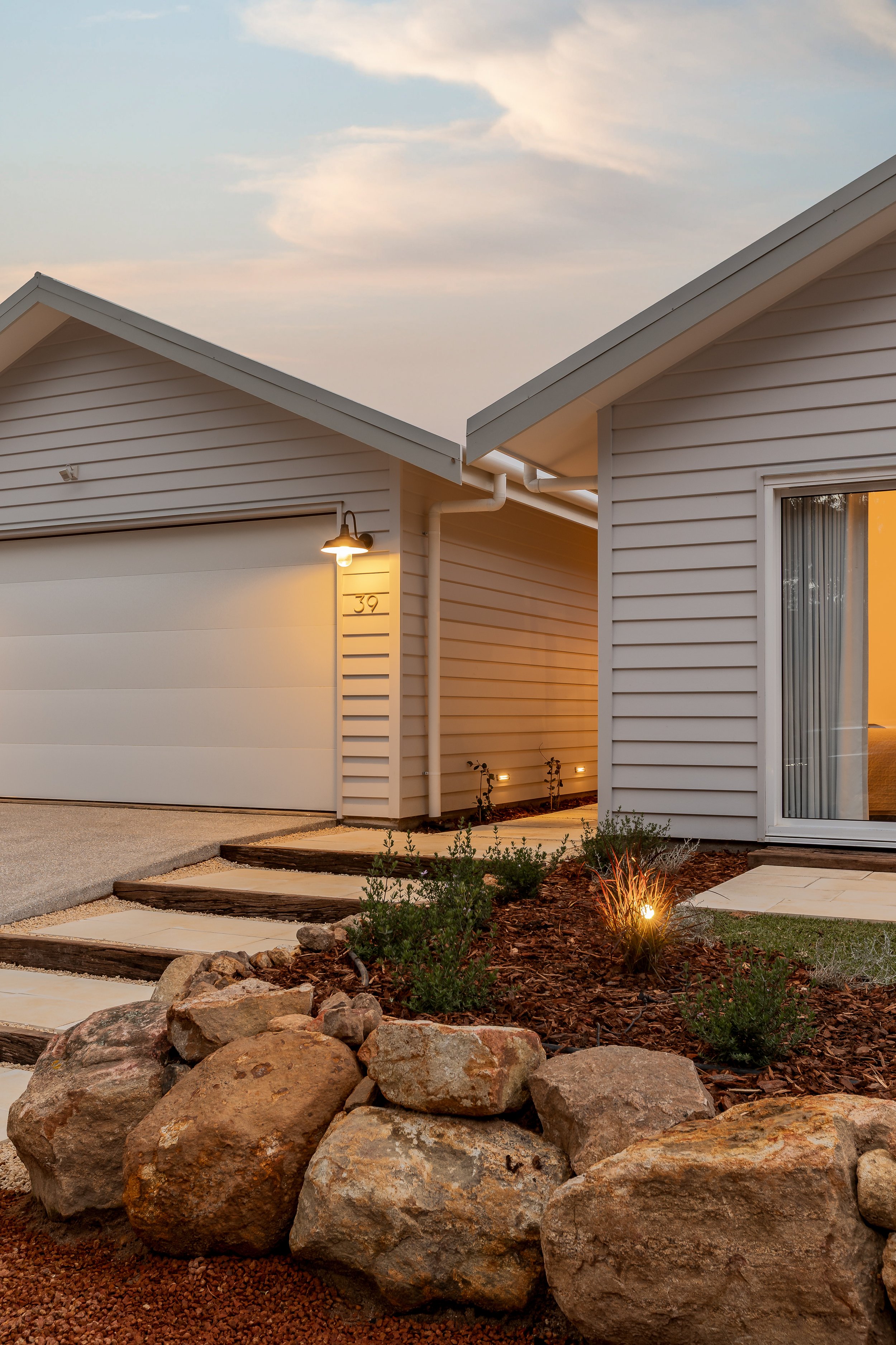 Cowaramup, Western Australia. The front exterior of a modern house with a garage door, white siding, a small garden with rocks, mulch, and small plants, and outdoor lighting at dusk. The house number '39' is visible. Real Estate Photography