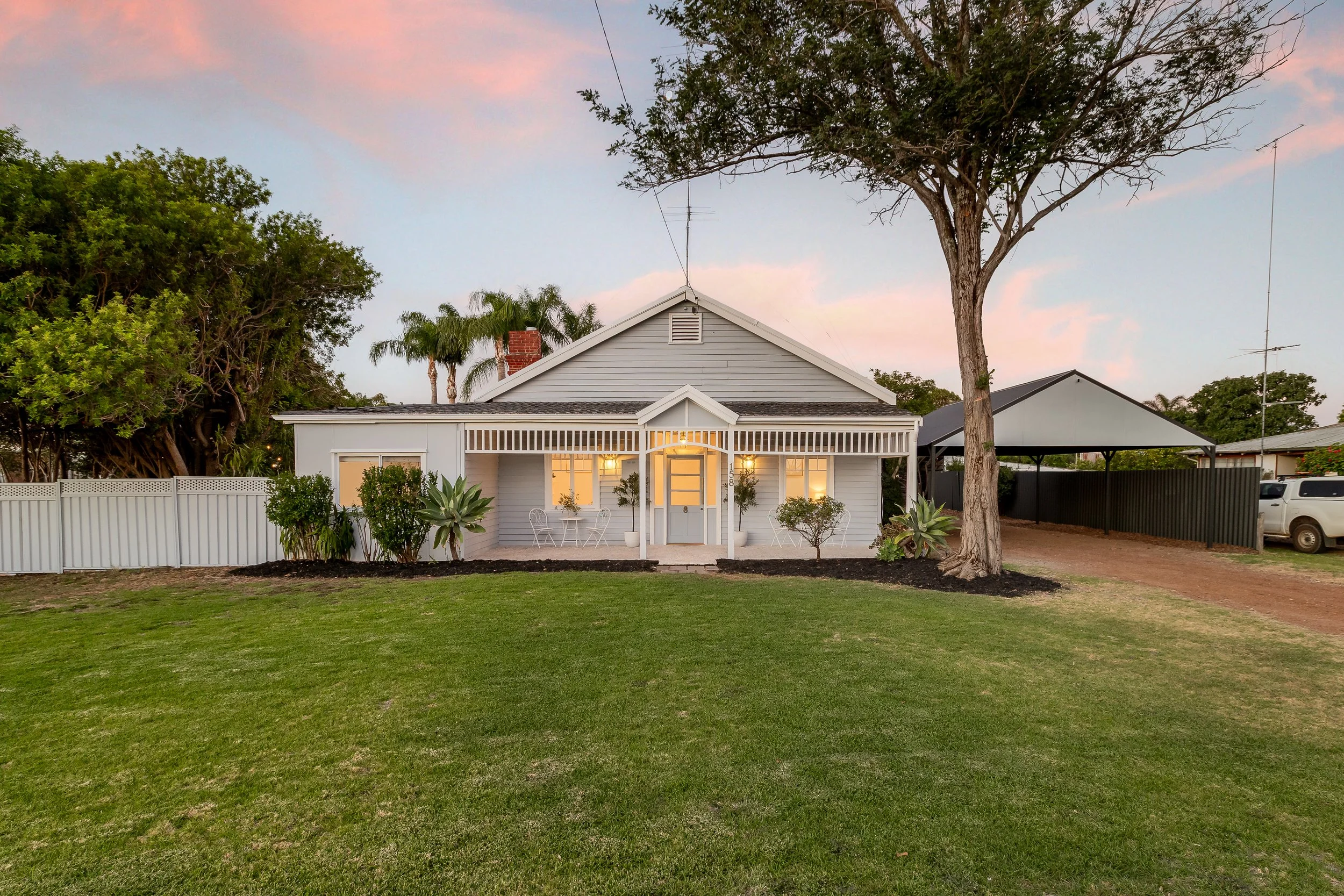 Harvey, Western Australia. A white house with a front porch illuminated by warm lighting, surrounded by green lawn and plants, with a large tree in front and neighboring houses and trees in the background at sunset. Real Estate Photography