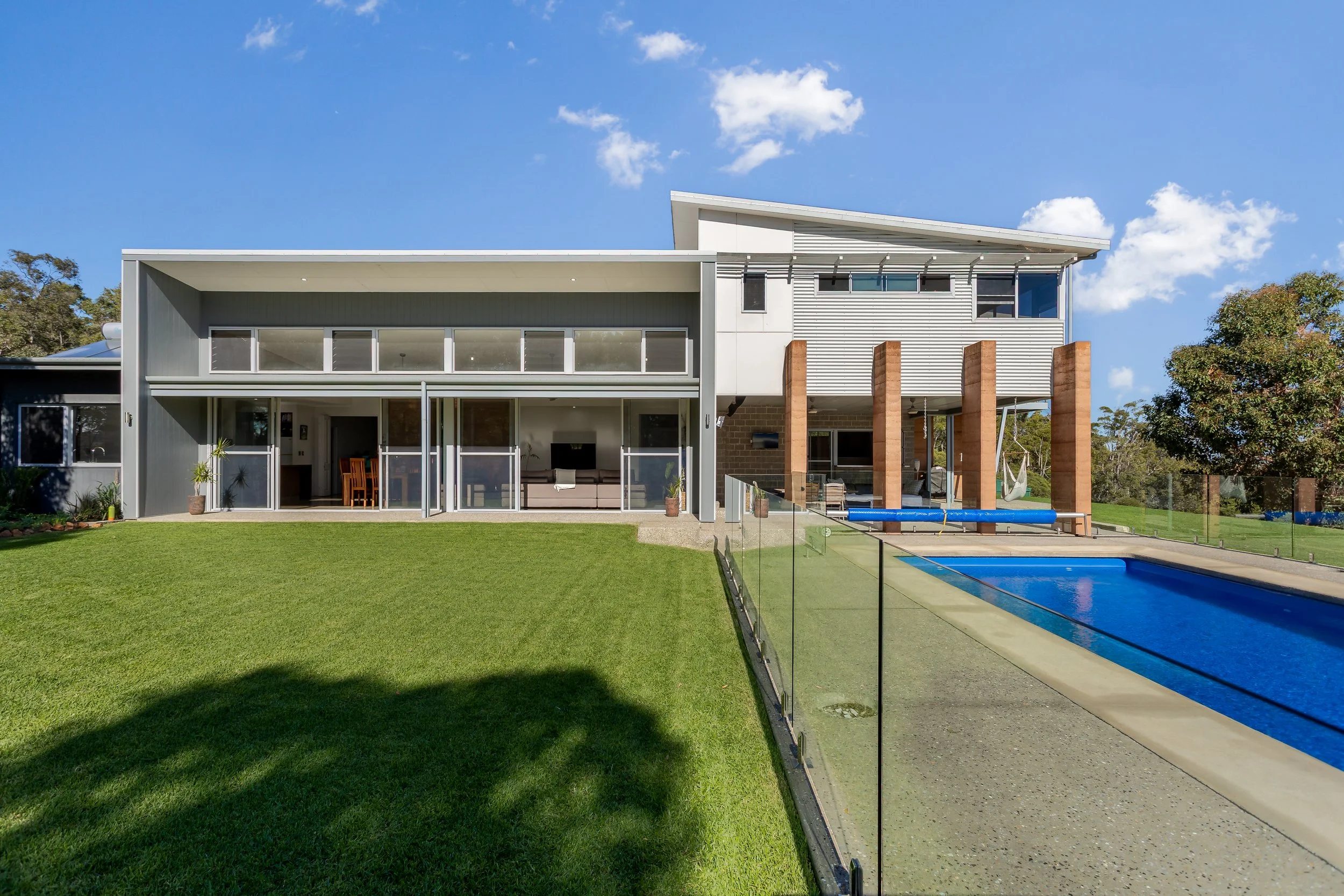 Crooked Brook, Western Australia. Modern two-story house with a backyard, swimming pool, green lawn, and trees under a blue sky with clouds. Real Estate Photography.