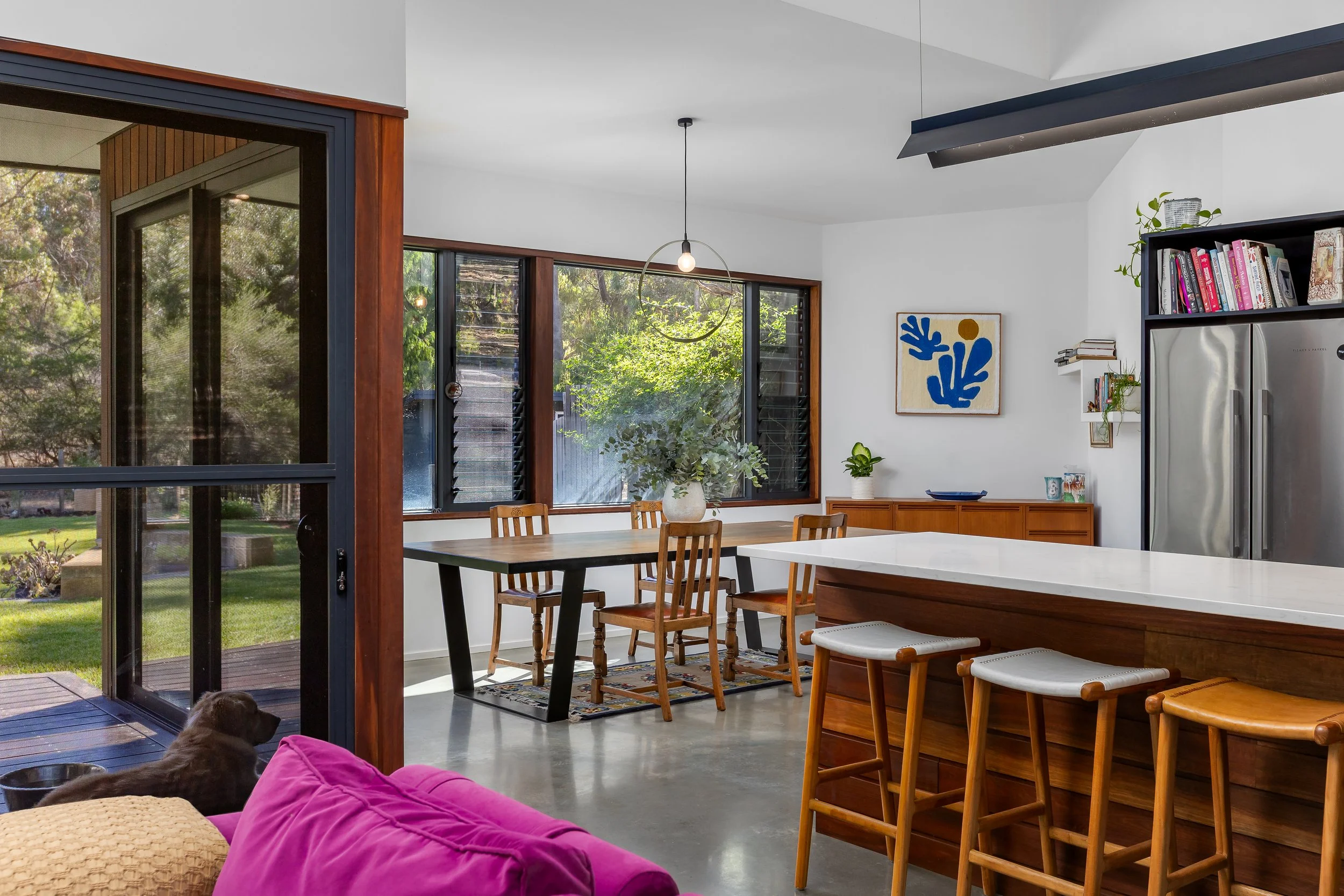 Yallingup, Western Australia. Modern living room with a large window and wood accents, showing a view of the outdoors with trees; includes a dining area, artwork on the wall, plants, and a kitchen. Real Estate Photography.