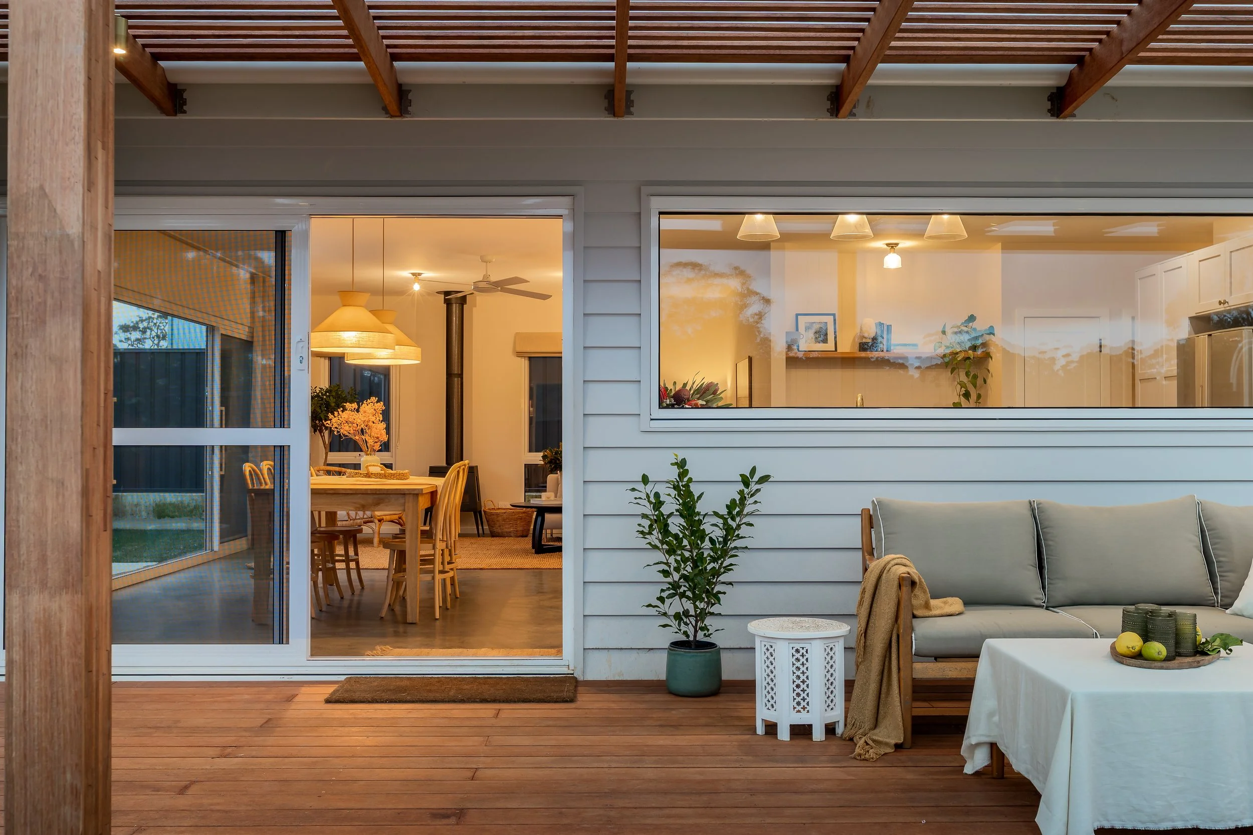 Margaret River, Western Australia. View of a house interior through large glass sliding doors, showing a dining area and a living room with a sofa and decorative plants, with warm ambient lighting. Real Estate Photography