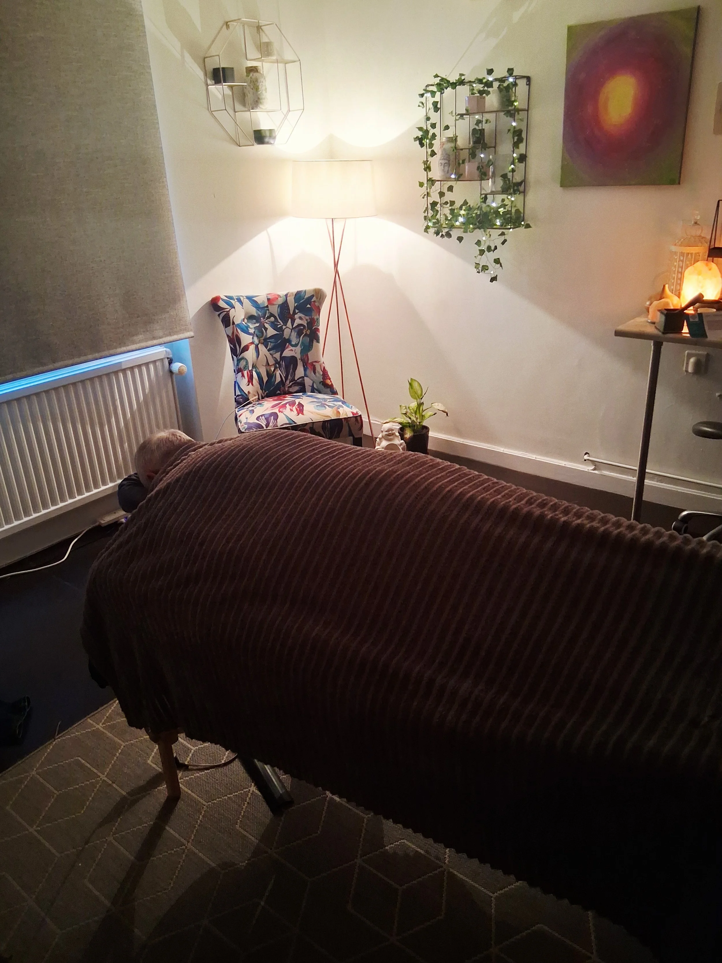 A cozy living room with a person lying face down on a table, a patterned chair, a lamp, shelves with plants and decorative items, a colorful painting, a Himalayan salt lamp, and a window with beige curtain.