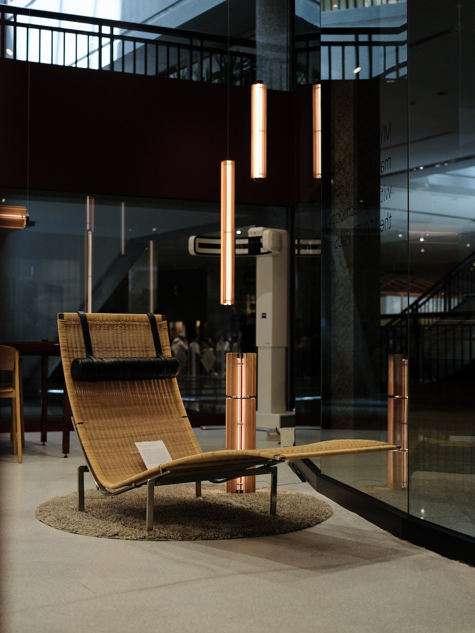 Modern interior display with a woven lounge chair on a circular rug, illuminated by vertical copper-colored pendant lights, with reflective glass walls and a partial view of an air conditioning unit in the background.