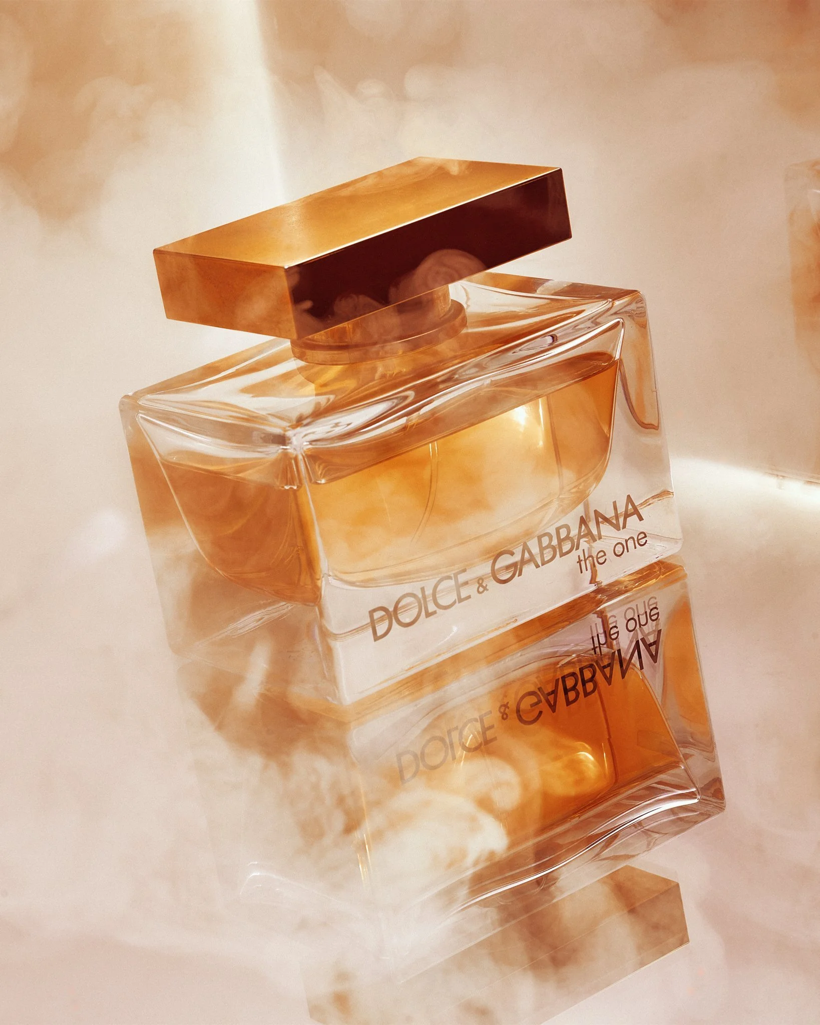 Dolce & Gabbana "The One" perfume bottle with golden liquid on a reflective surface surrounded by mist.