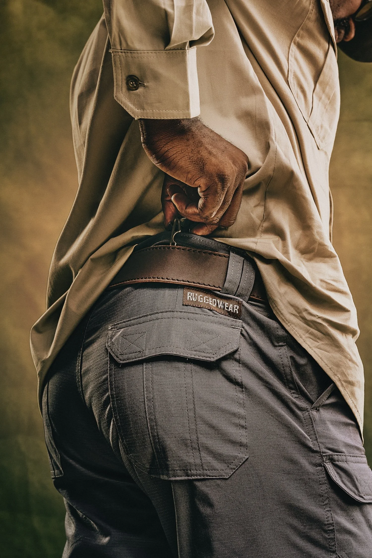 Close-up of a person wearing a beige shirt and dark pants with a "Rugged Wear" label, adjusting their belt.
