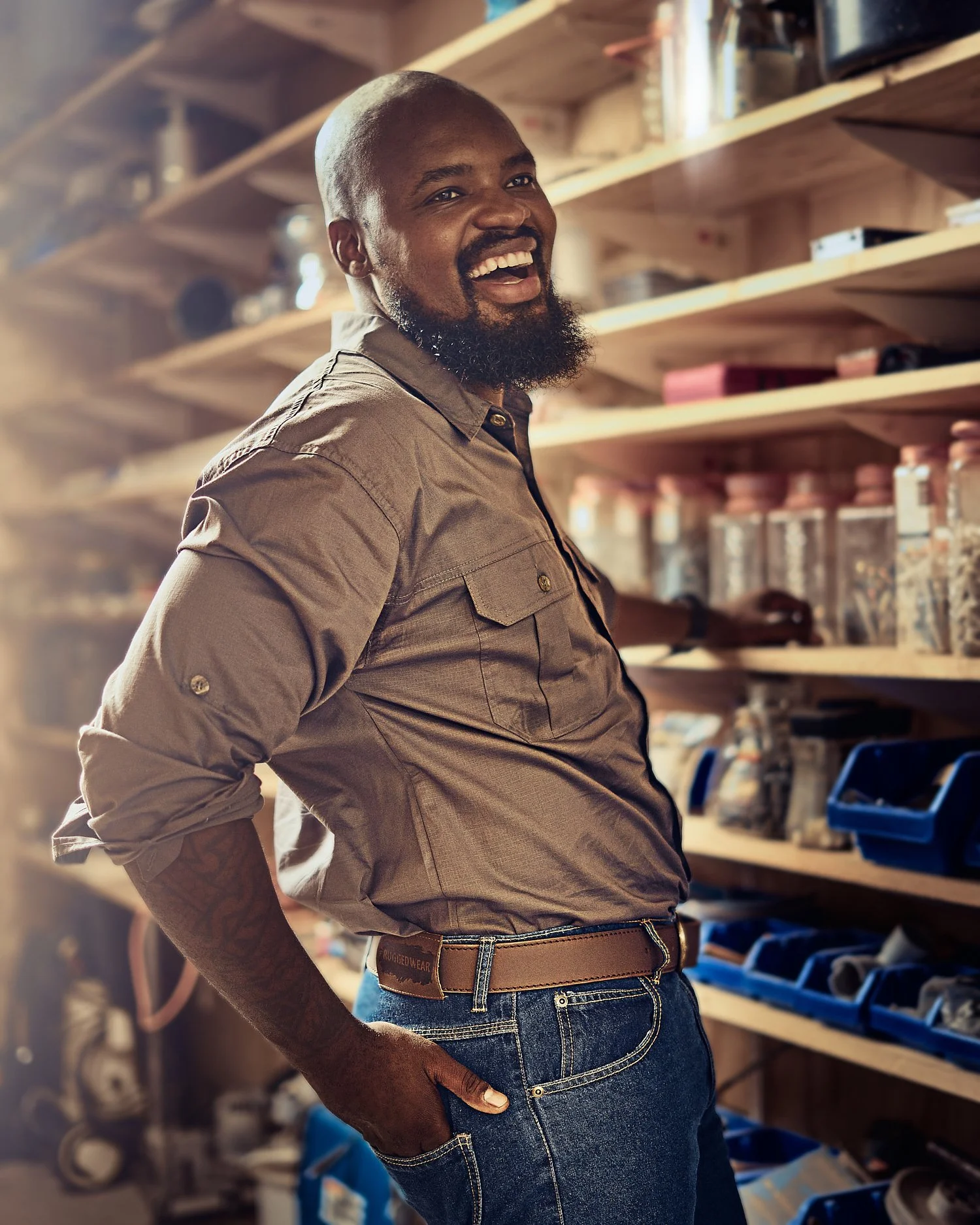 Smiling person in a workshop, wearing a brown shirt and jeans, with shelves of tools in the background.