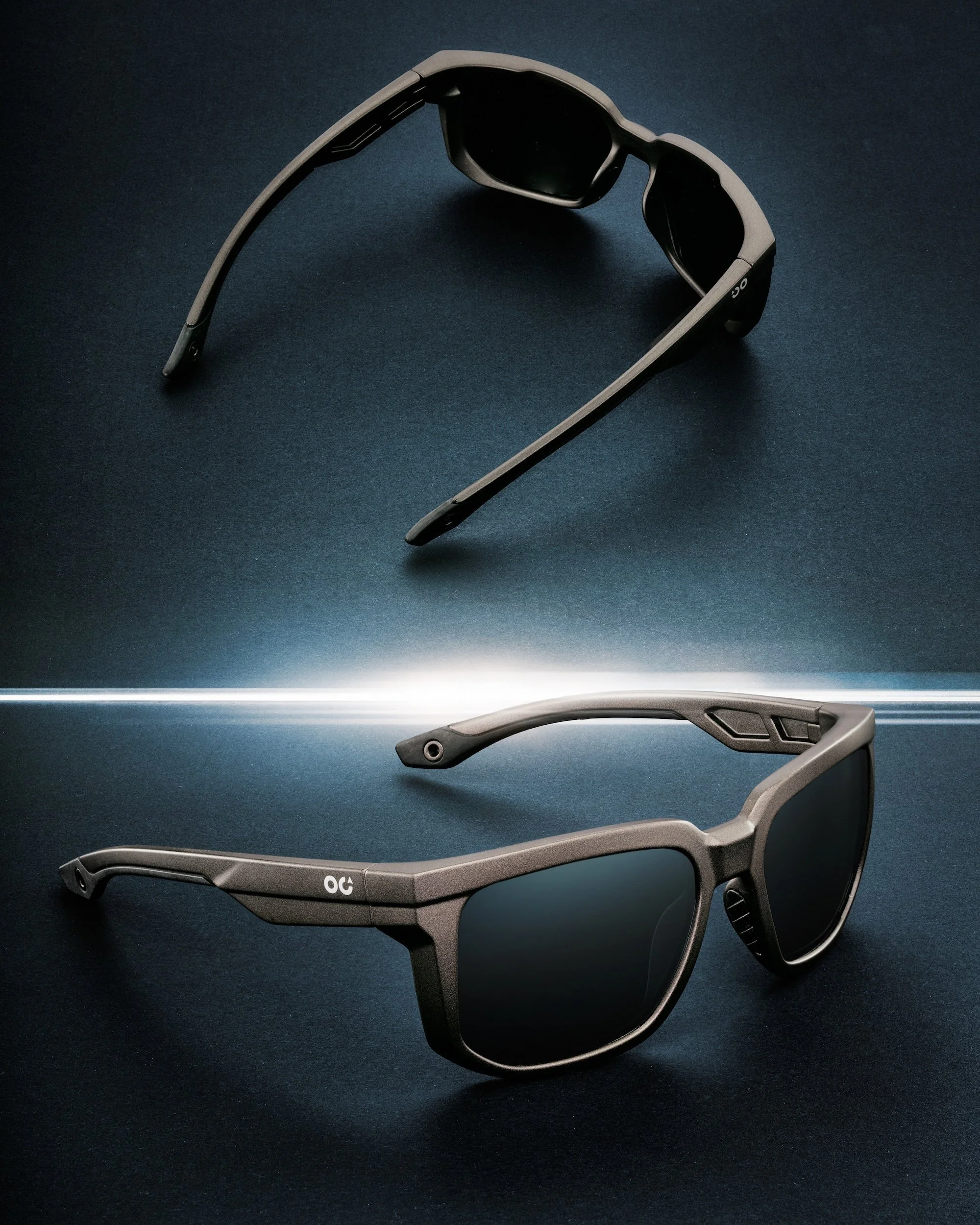 Two pairs of sporty sunglasses with dark lenses on a dark background, one positioned above the other with a light source separating them.