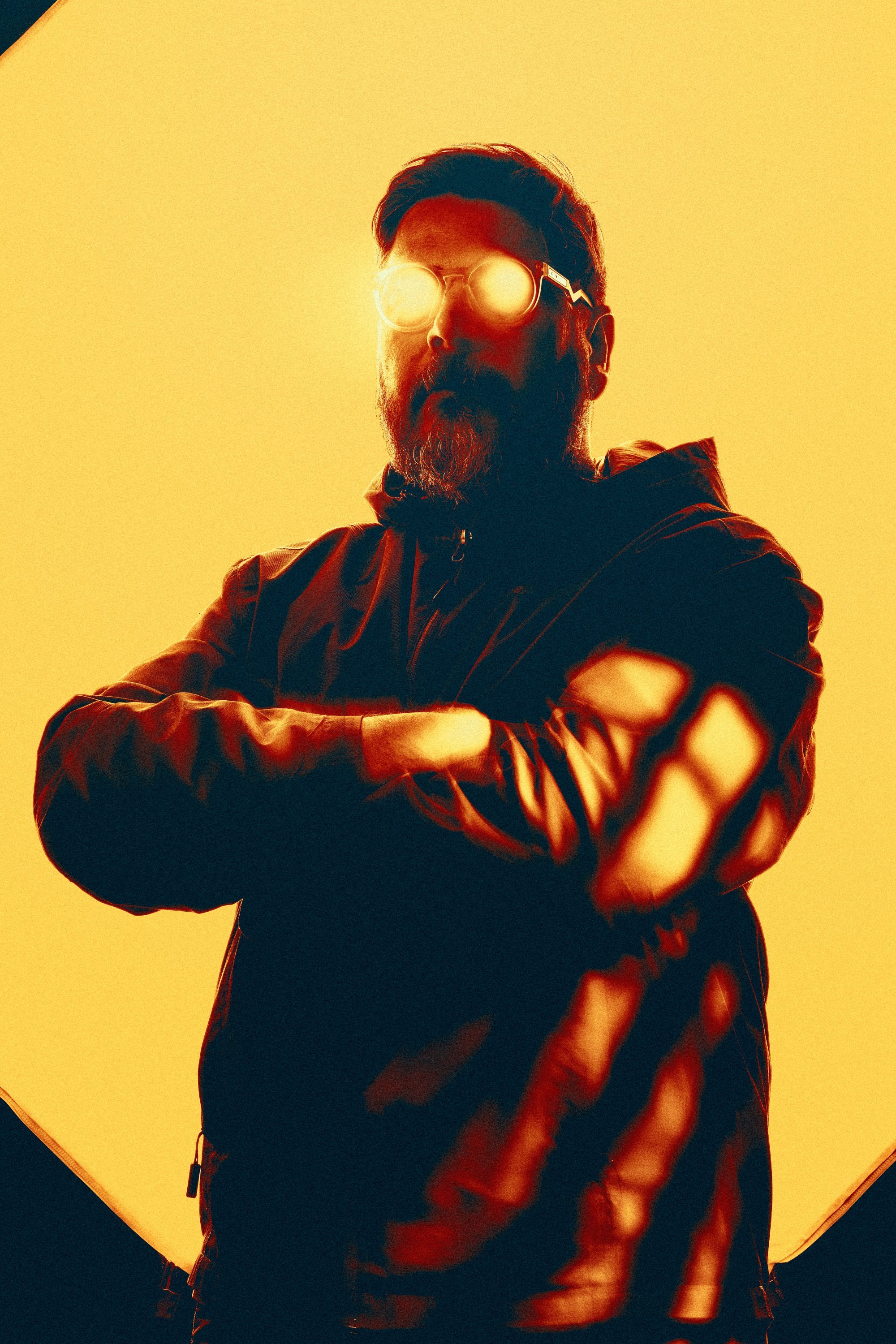 Stylized photo of a man with glasses emitting light, wearing a jacket, standing with arms crossed against a bright yellow background.