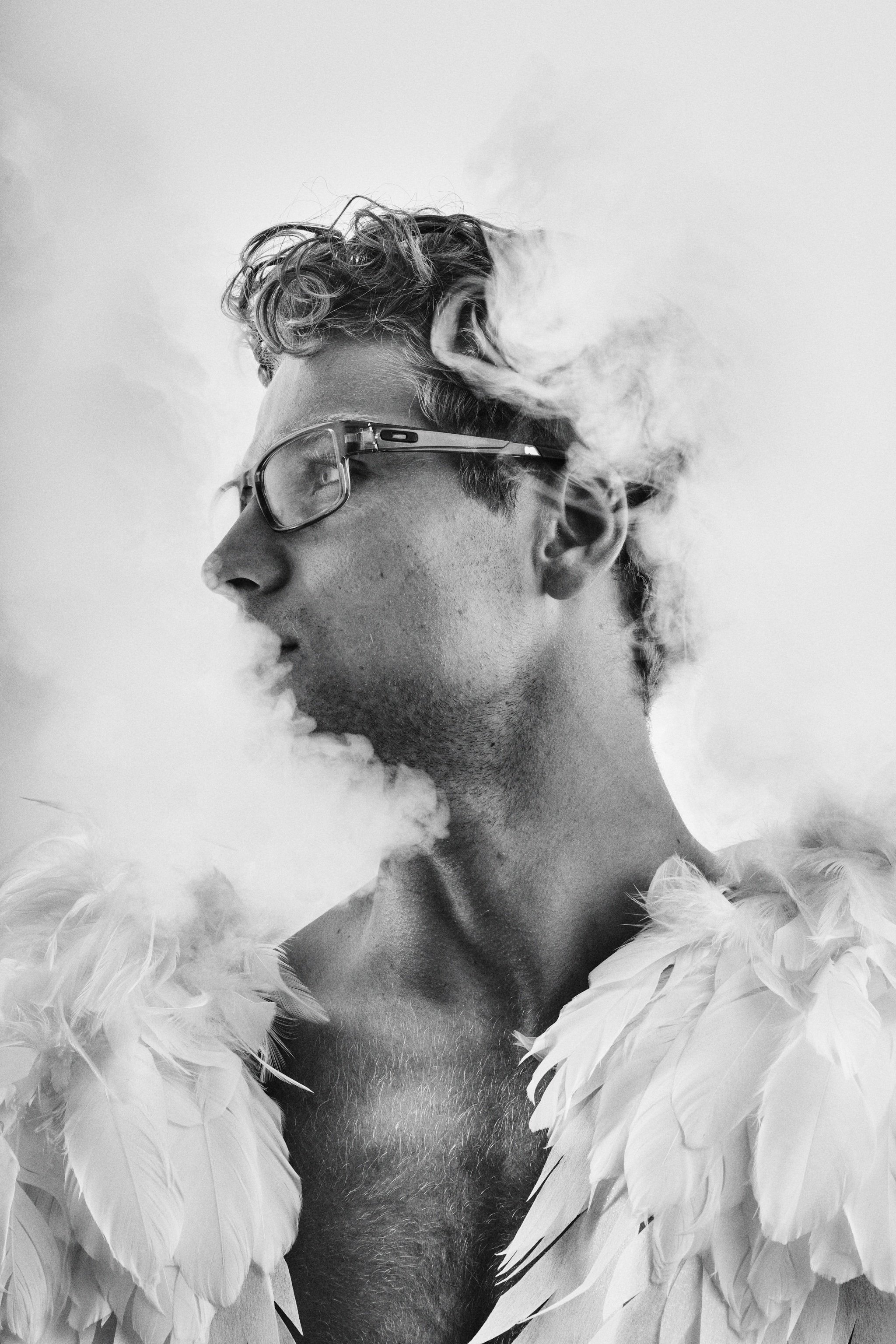 Black and white portrait of a man with glasses surrounded by smoke, wearing feather wings.