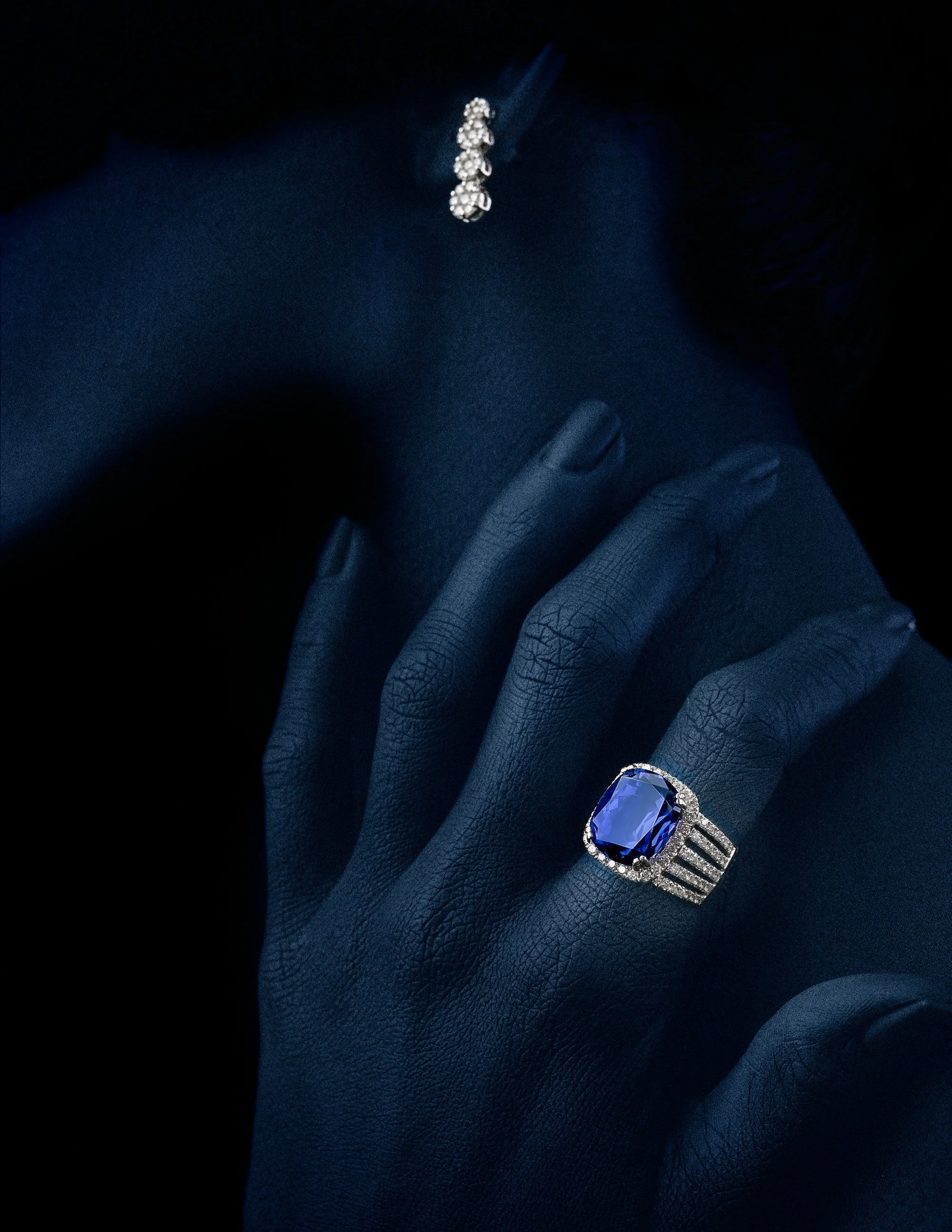 Close-up of a hand wearing a Tanazinte large ring with a blue gemstone and surrounding diamonds, against a dark background with part of a necklace visible.
