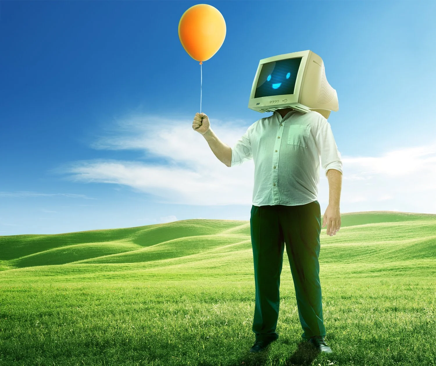 A person standing in a grassy field with a clear blue sky, wearing a vintage computer monitor as a head and holding an orange balloon.